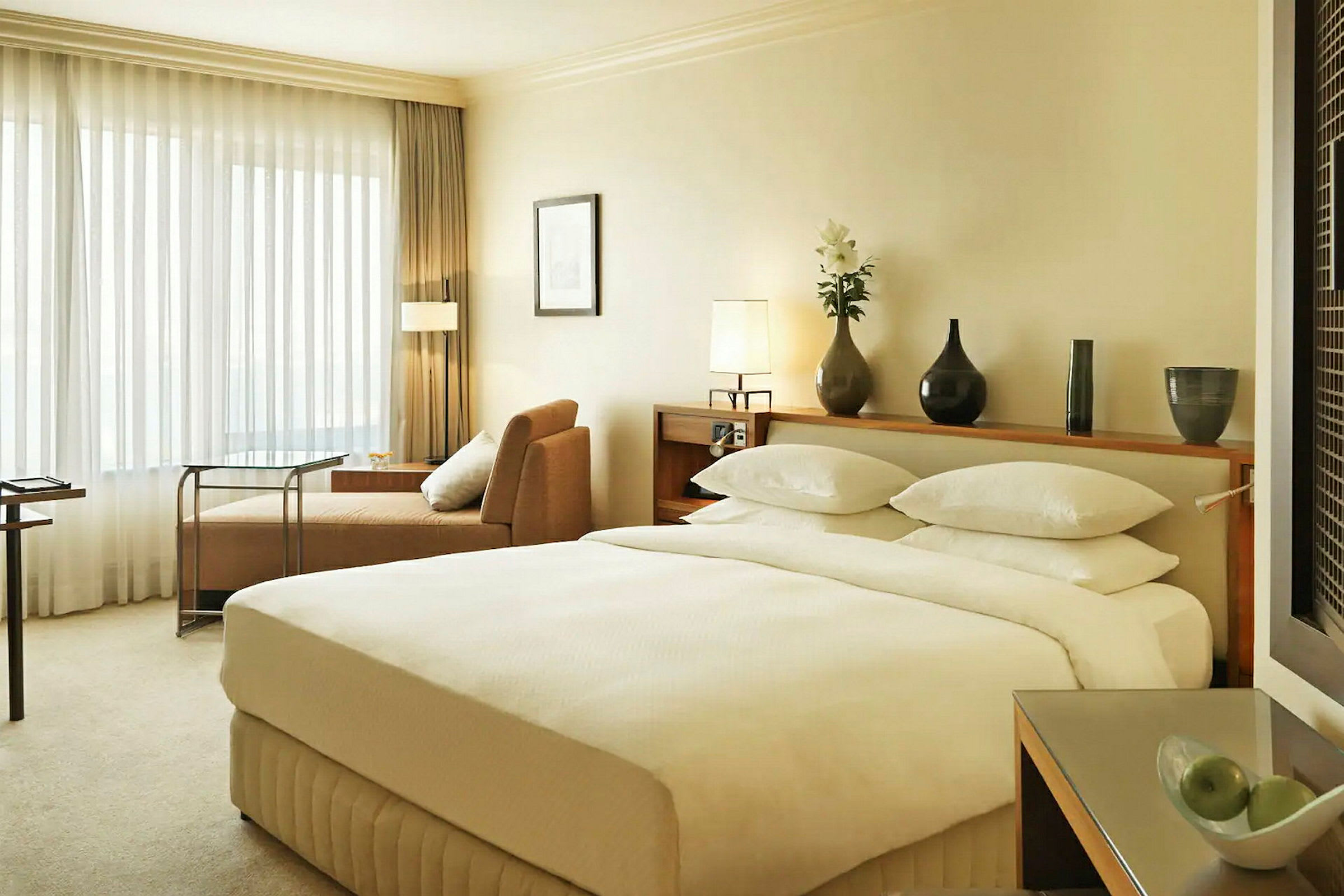 Soveværelse i hotelværelse med en stor dobbeltseng, beige sofa og et bord nær et vindue med gennemsigtige gardiner. (AI-genereret tekst)