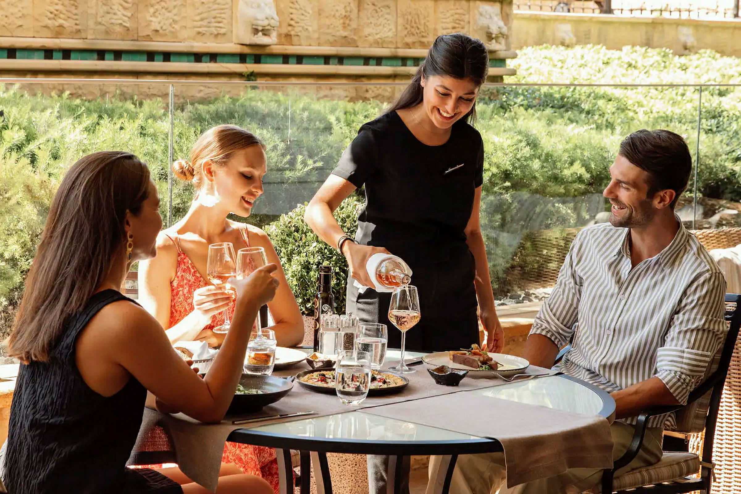 En tjener serverer drikke til gæster, der sidder ved et bord på en udendørsrestaurant. (AI-genereret tekst)
