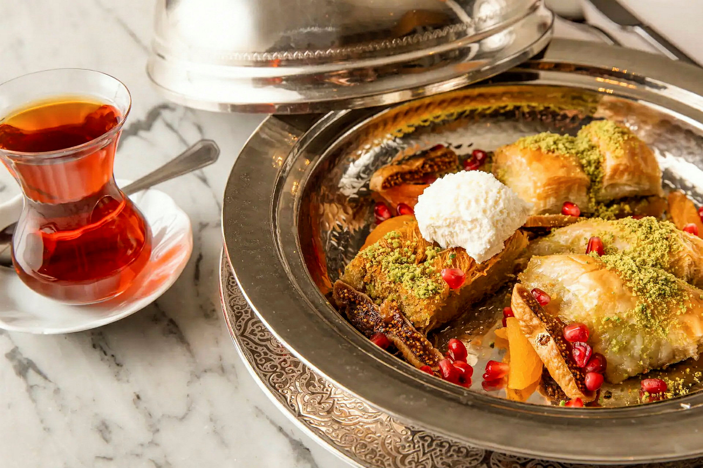 En tallerken med dessert bestående af baklava, pistacienødder, granatæblekerner og flødeskum, ved siden af et glas te. (AI-genereret tekst)
