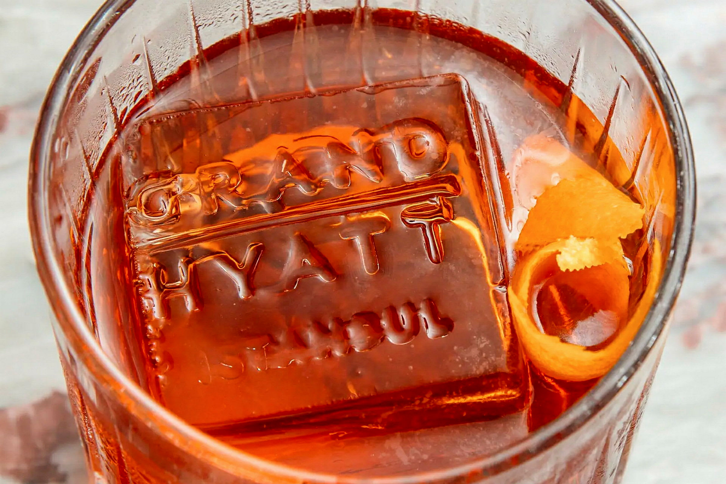 Et glas med en orange drik og en isterning præget med teksten 'Grand Hyatt Istanbul'. (AI-genereret tekst)