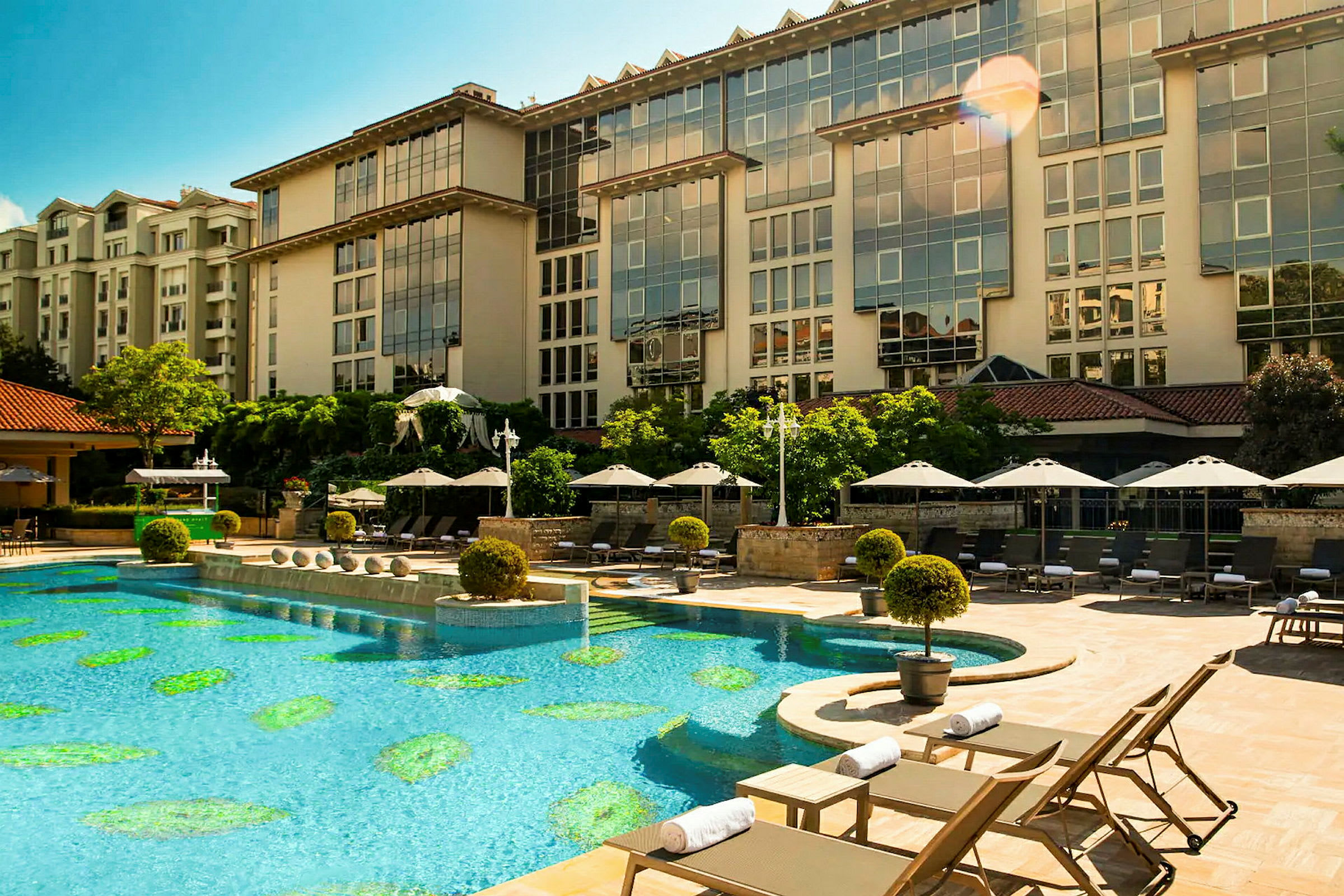 En udendørs pool ved et hotel omgivet af solsenge og parasoller. Hotelbygningen ses i baggrunden. (AI-genereret tekst)