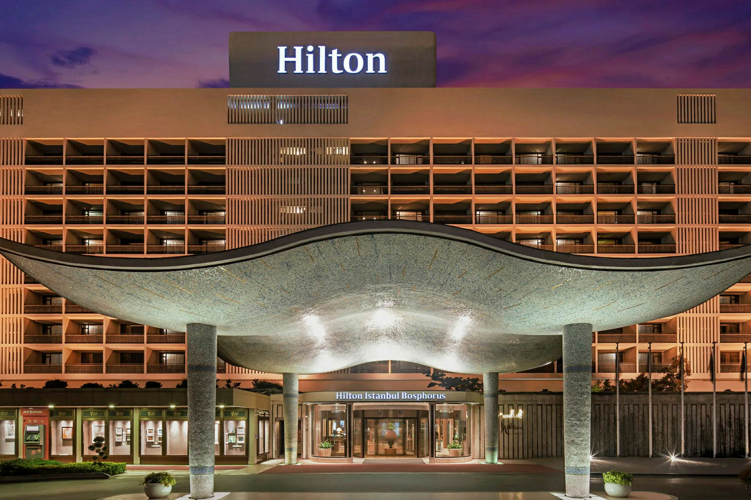 Hotellets facade med indgang under et hvælvet tag, lygtepæle og et oplyst skilt med teksten 'Hilton'. (AI-genereret tekst)