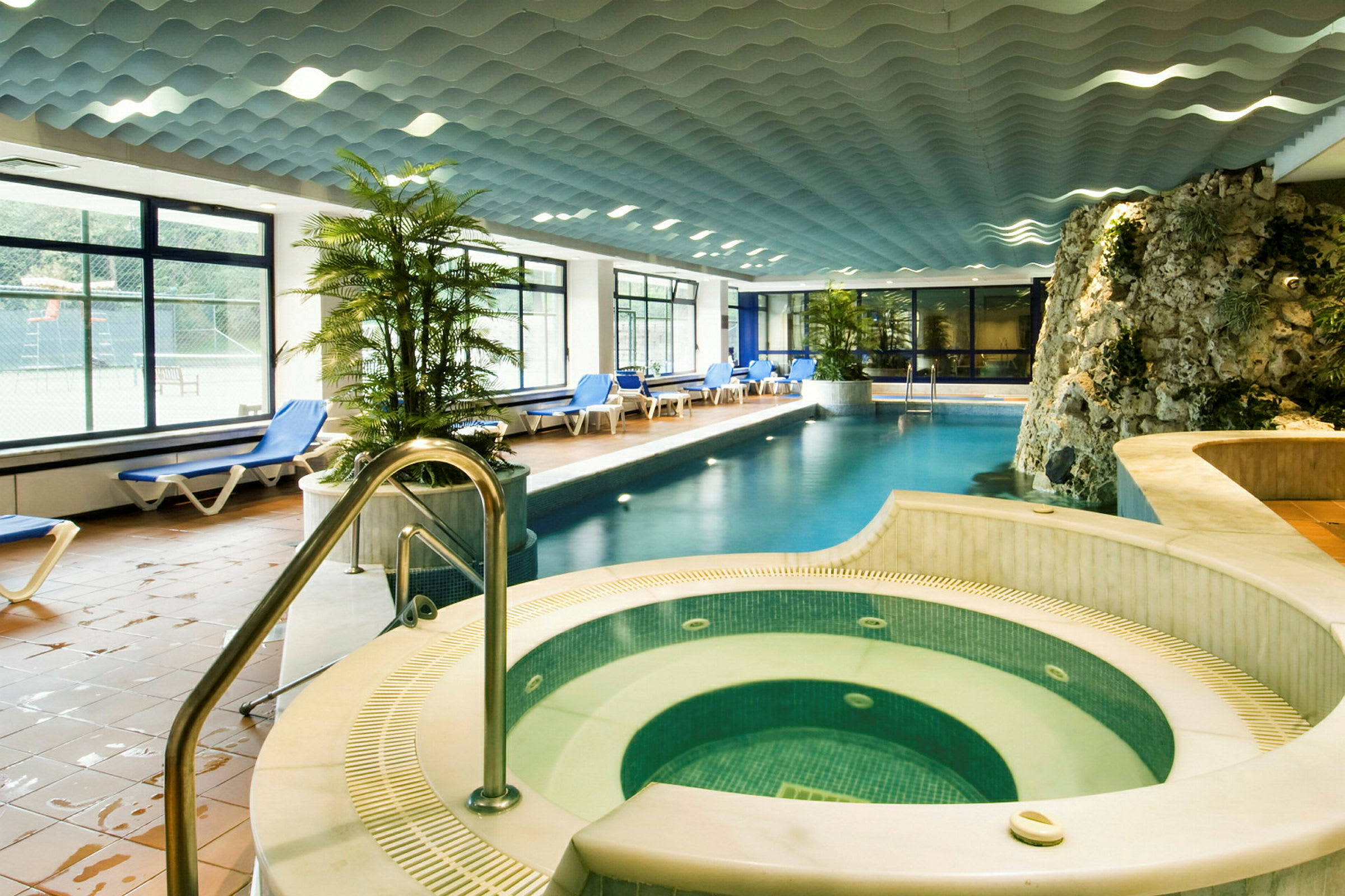 Et indendørs swimmingpool med solsenge langs kanterne og grønne planter som dekoration. I forgrunden er et spabad omgivet af mosaik. Loftet har et bølgemønster. (AI-genereret tekst)