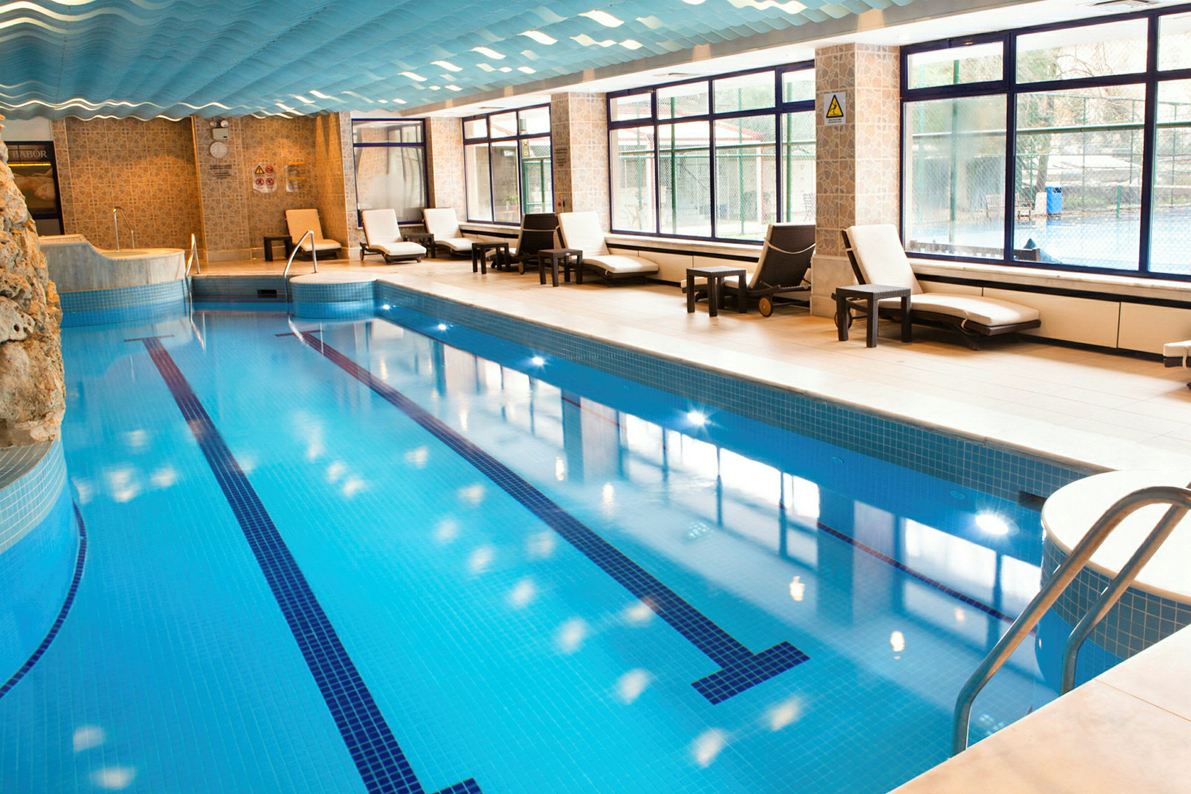Billede af en indendørs swimmingpool med liggestole og vinduer, der lukker naturligt lys ind. (AI-genereret tekst)