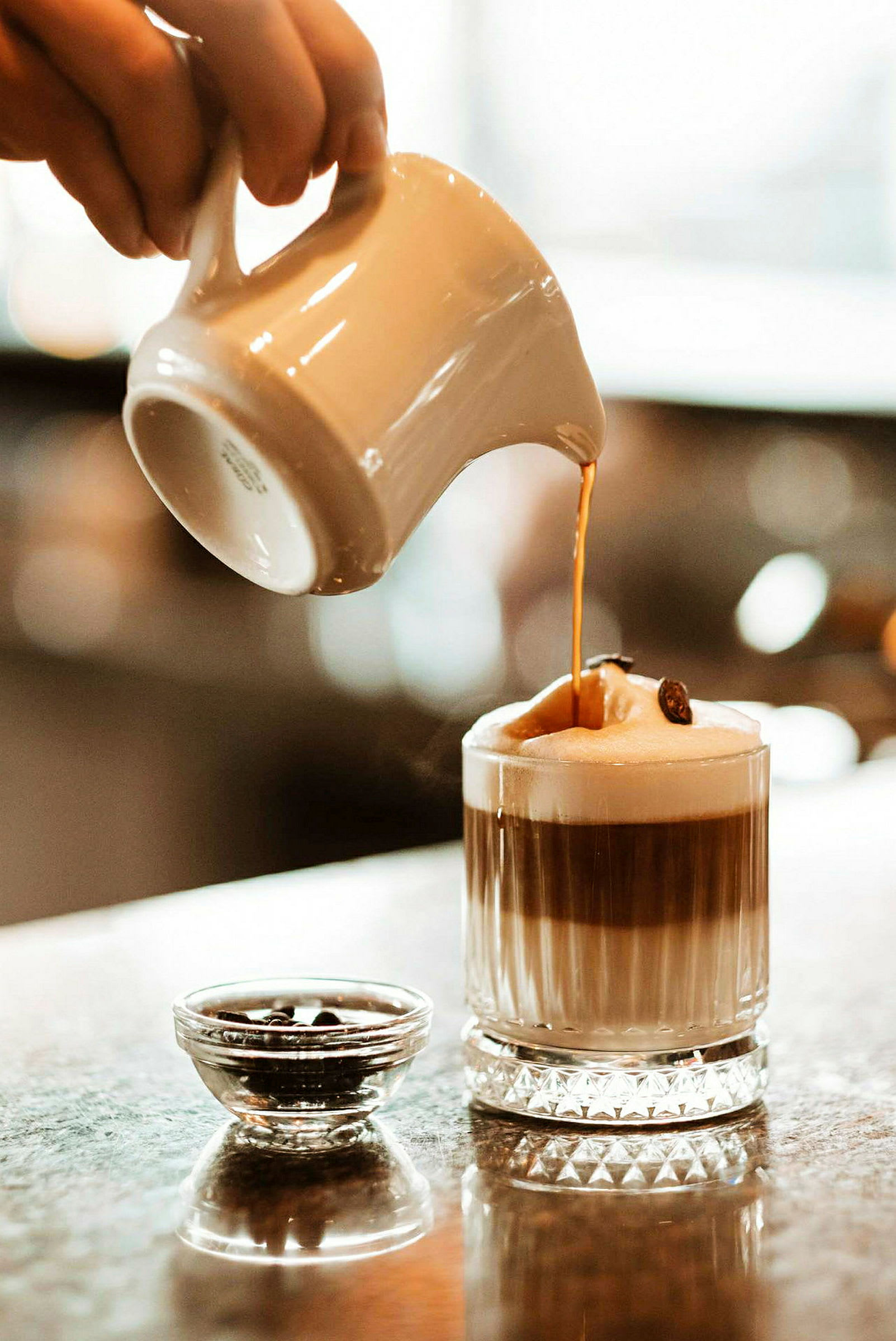 En hånd hælder en varm drik, muligvis kaffe, fra en kande ned i et glas med skum på toppen. Ved siden af glasset står en lille skål med kaffebønner. (AI-genereret tekst)