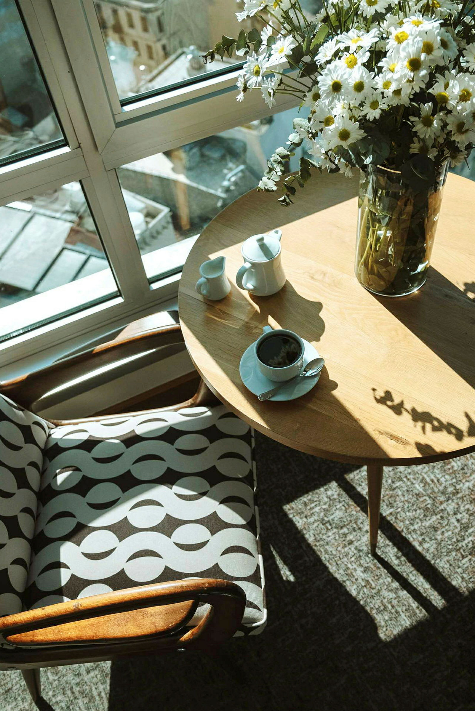 Et bord med en kaffekop, sukkerbit og en mælkekande ved siden af en stol med mønstret stof, placeret foran et stort vindue med udsigt. (AI-genereret tekst)