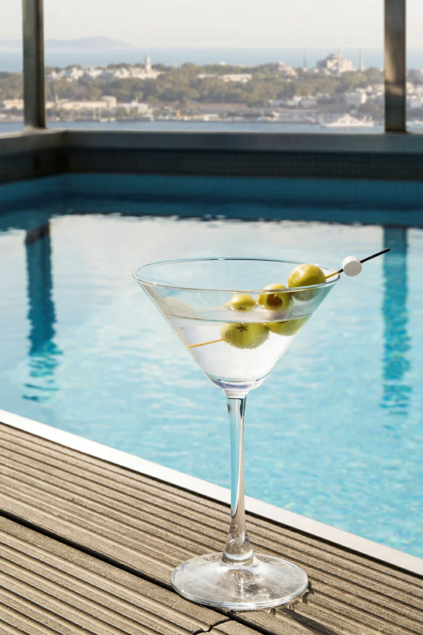 Et glas med en cocktail og oliven på en træflade ved siden af en pool, med udsigt over et bylandskab. (AI-genereret tekst)