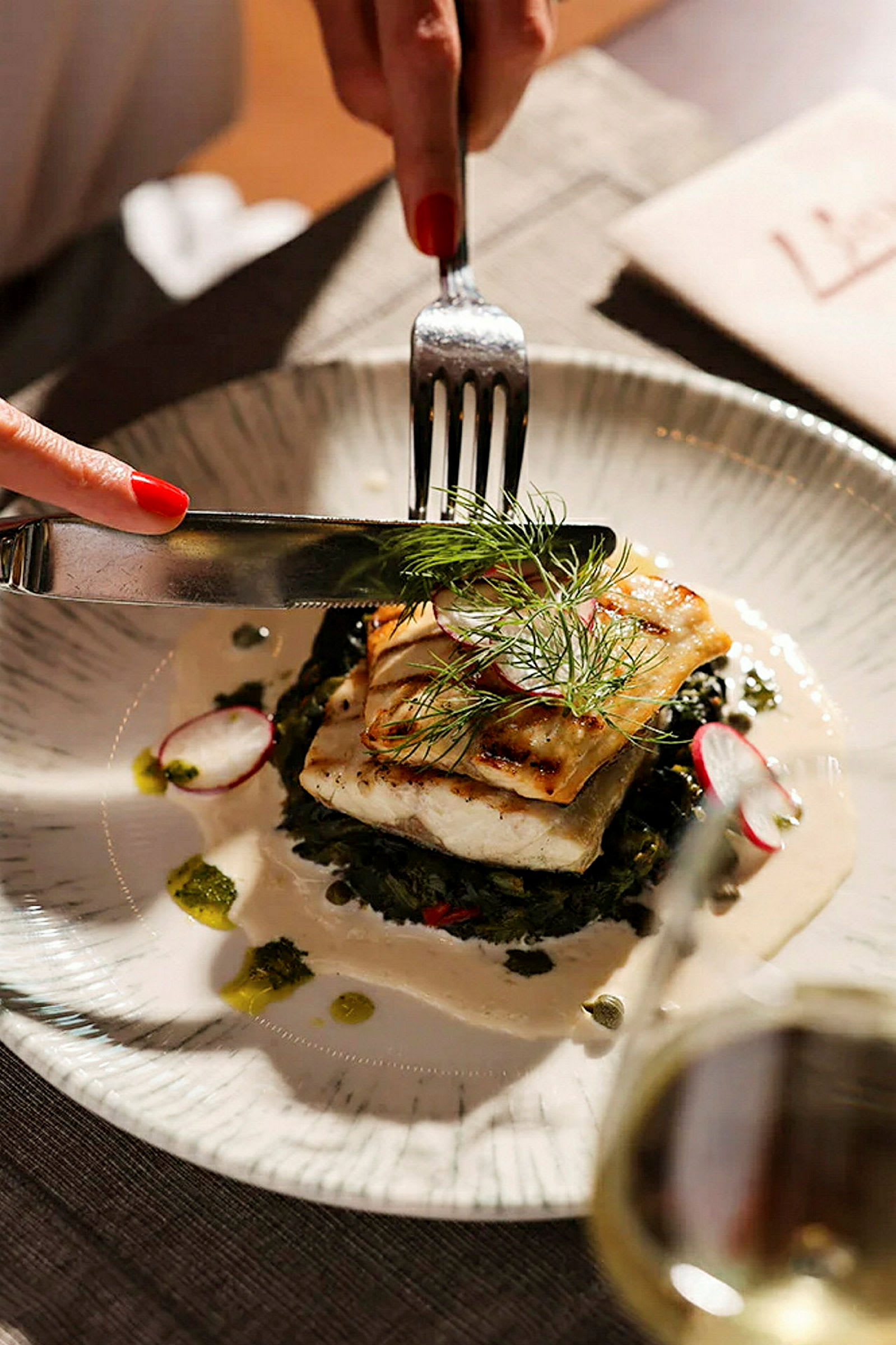 En tallerken med et måltid bestående af grillet fisk, serveret med grøntsager og en sauce. Bestik bruges på maden. (AI-genereret tekst)