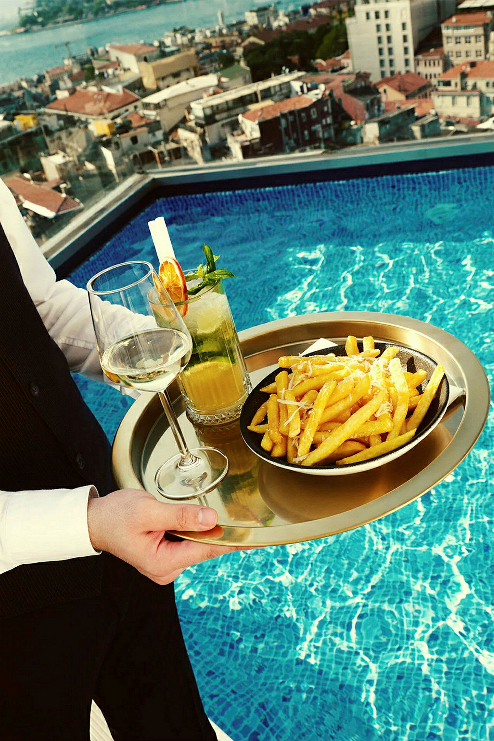 En serveringsbakke med pommes frites på en tallerken, et glas hvidvin og en cocktail er placeret ved en pool med udsigt over en by. (AI-genereret tekst)