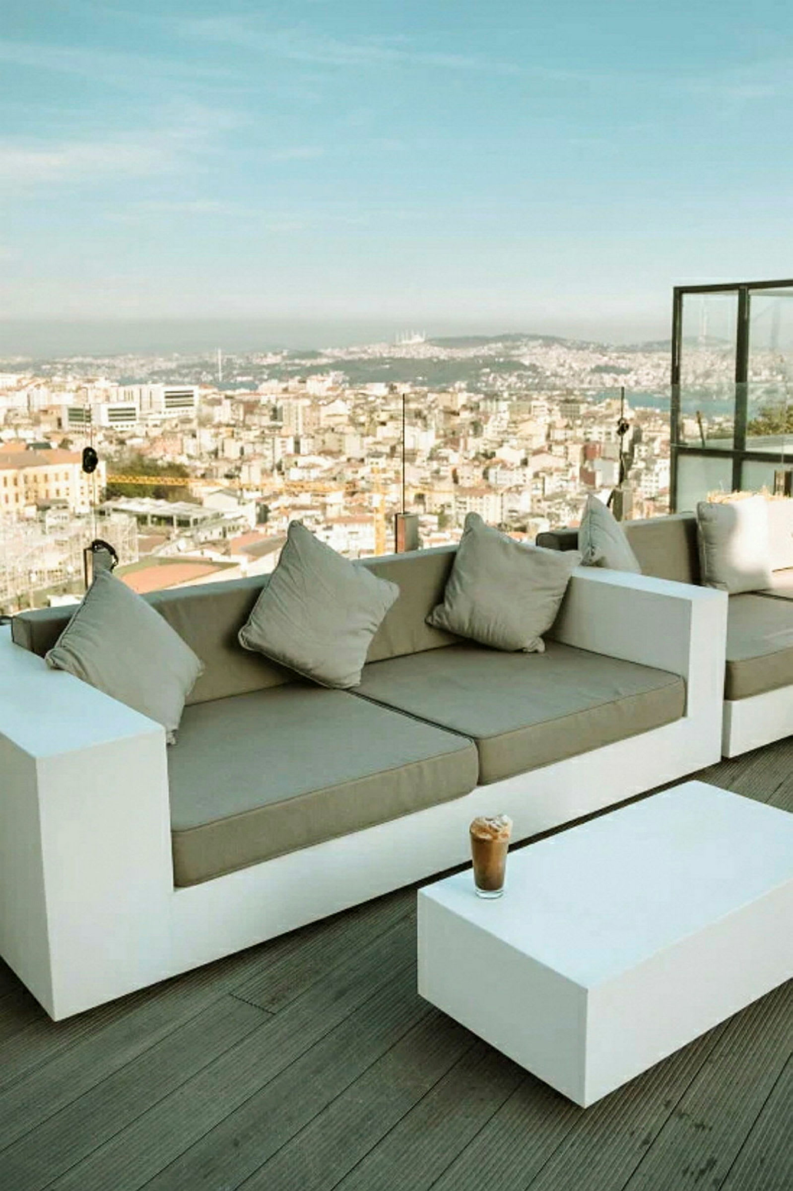 En moderne terrasse med hvide sofaer og et lavt bord, med udsigt over en by og en klar himmel. (AI-genereret tekst)
