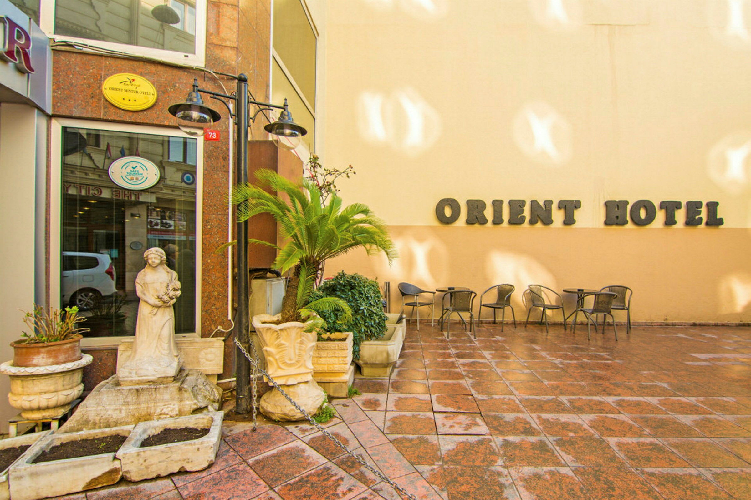 En bygning med skiltet "Orient Hotel" på en lys væg, med en lille terrasse dekoreret med stole, borde, blomsterpotter og en statue. (AI-genereret tekst)