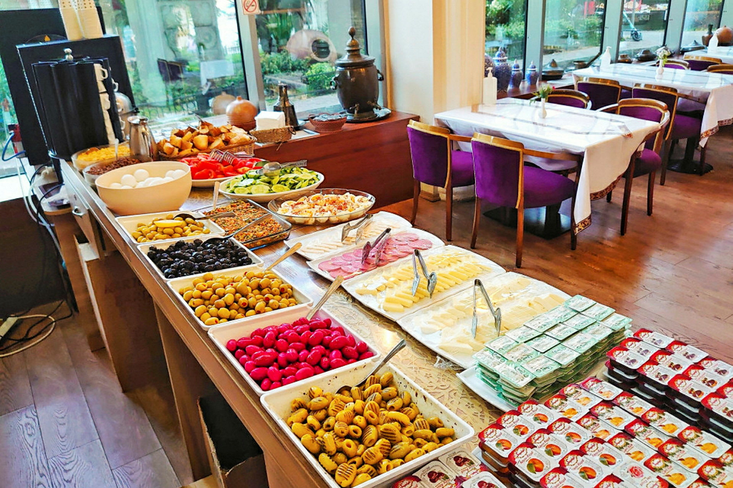 Buffet med forskellige retter, skåle og tallerkener fyldt med grøntsager, oste og pålæg. I baggrunden ses spiseområde med dækkede borde og stole. (AI-genereret tekst)