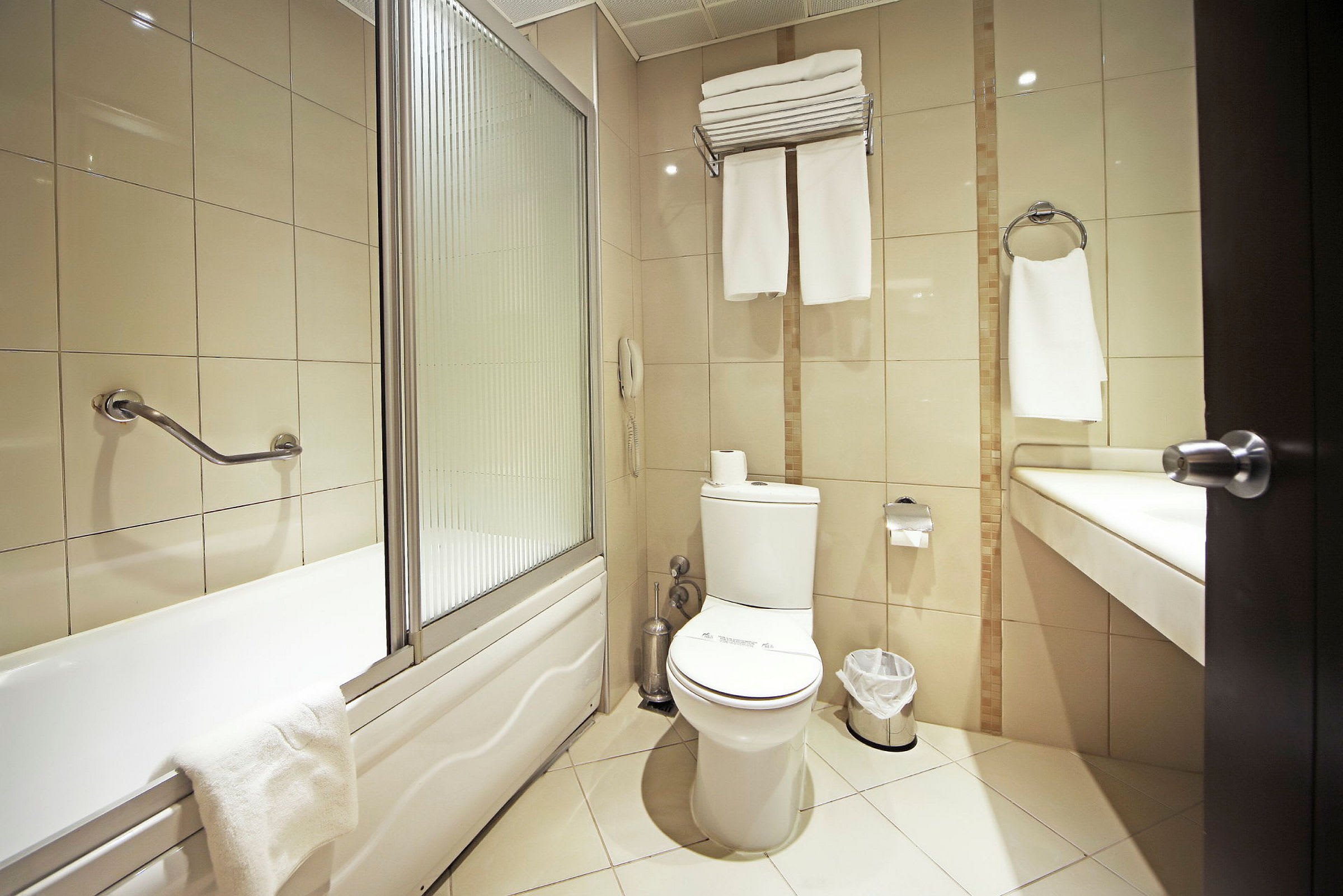 Et badeværelse med et badekar, toilet og håndvask. Væggene har beige fliser, og badeværelset er udstyret med håndklæder og en papirkurv. (AI-genereret tekst)