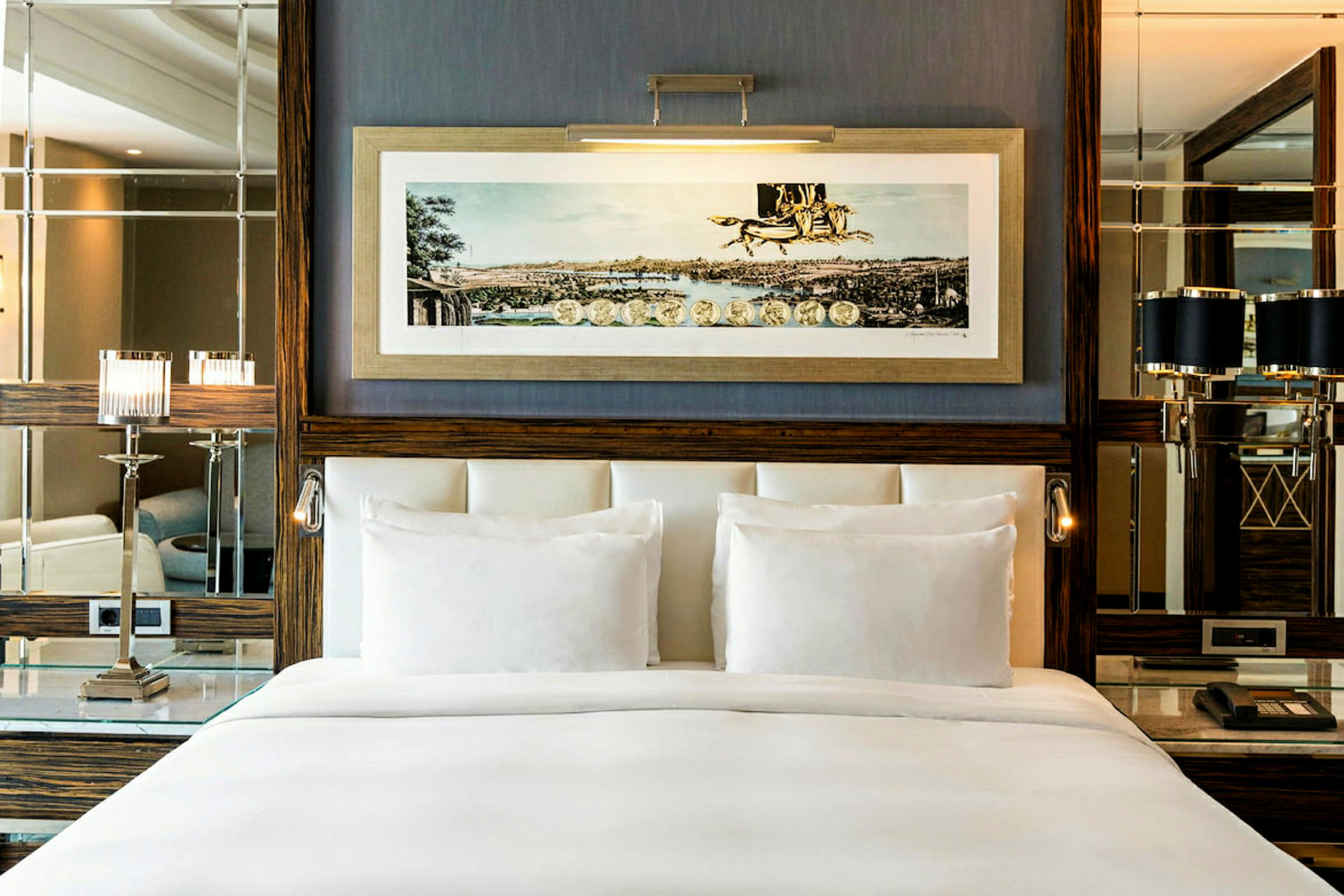 Et elegant hotelværelsesinteriør med en bred, hvidopredt seng foran en væg dekoreret med indrammet kunstbillede, spejle og elegante lamper. (AI-genereret tekst)