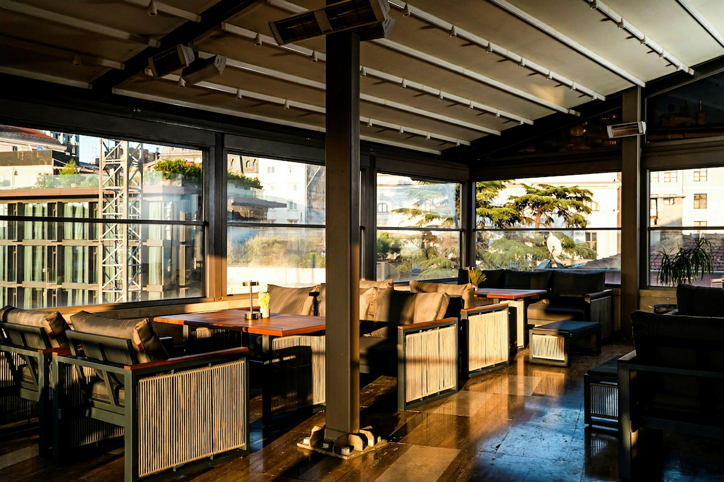 Billede af en elegant indrettet indendørs terrasse med møbler i mørkt træ og puder, med store vinduer og et loft for lysindfald. (AI-genereret tekst)