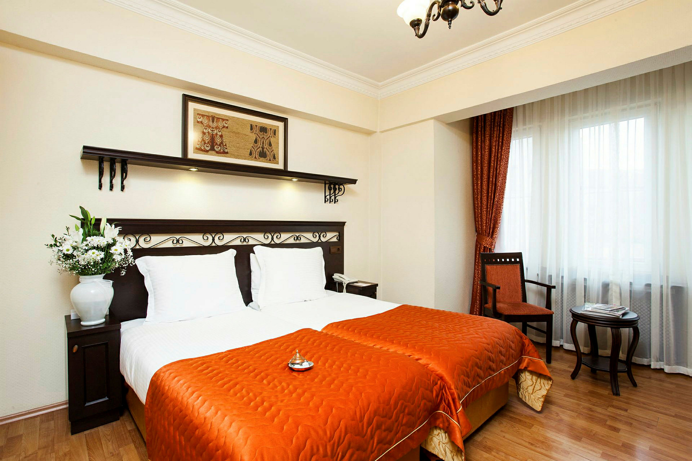 Et hotelværelse med to enkeltsenge dækket med orange sengetøj. Sengene har hvide puder og en mørk træ-sengegavl med dekorative detaljer. En blomsterkrukke står på et mørkt træbord ved siden af sengene. Vinduerne har hvide gardiner og orange draperier, og ved siden af er der en stol og et lille sidebord. (AI-genereret tekst)