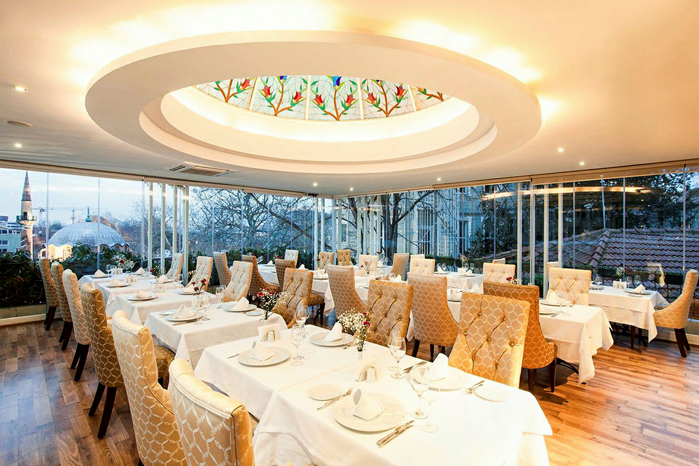 En elegant restaurant med dækkede borde og gyldne polstrede stole. Et stort vindue viser udsigten over en by med træer og bygninger. (AI-genereret tekst)