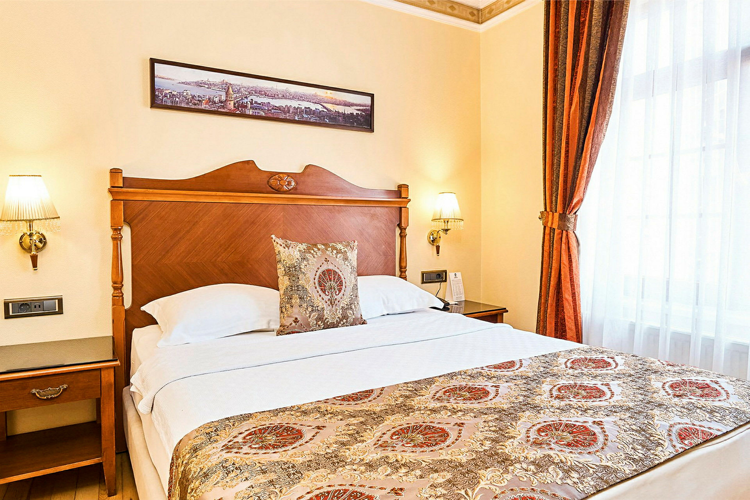 Hotelværelse med en dobbeltseng og elegante dekorationer. Sengens hovedgærde er af træ, og sengen har et dekoreret sengetæppe samt pyntepuder. På væggen hænger et panorama som kunst. (AI-genereret tekst)