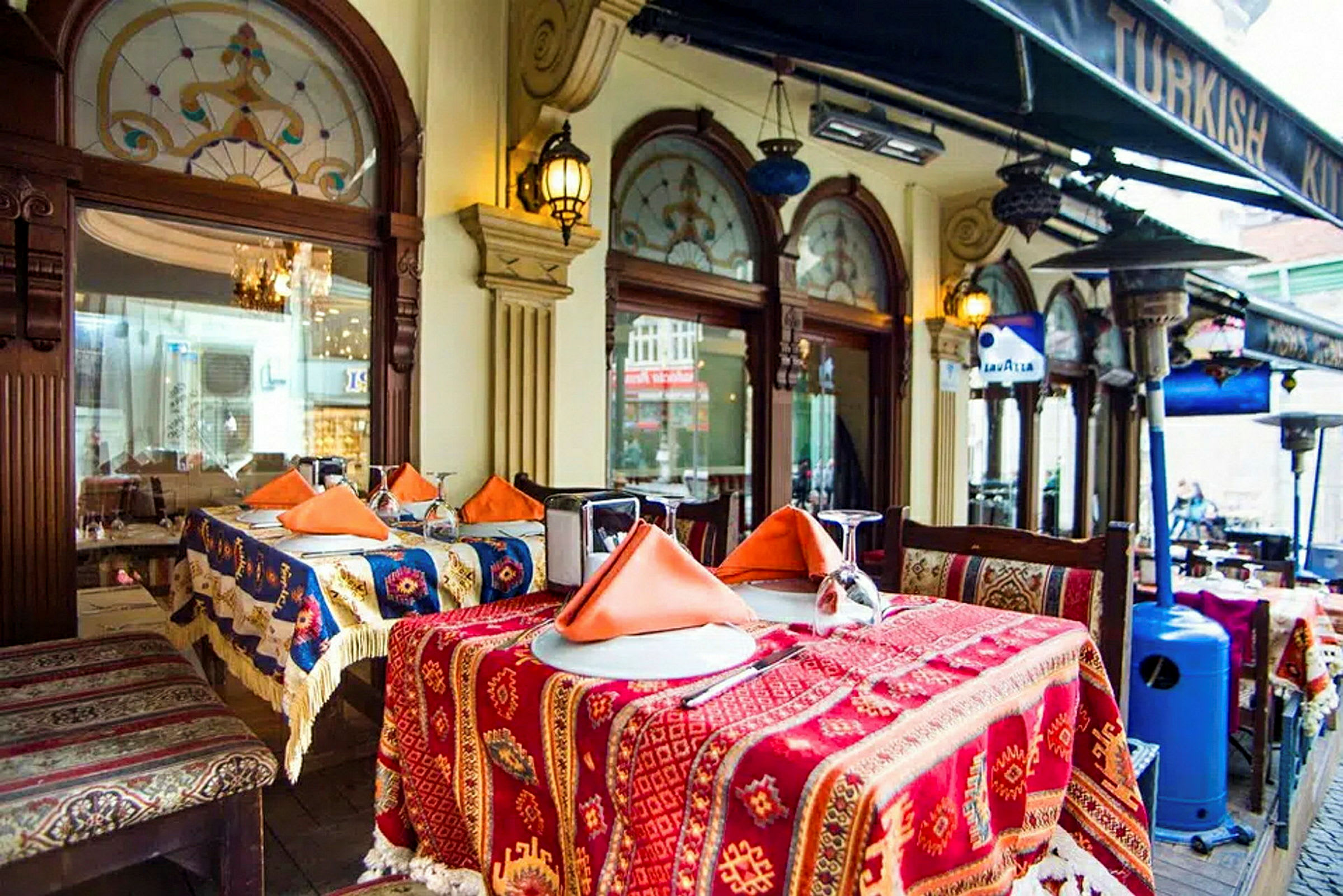 Borde dækket med farverige duge og orange servietter uden for en restaurant med dekorerede vinduer og et tag under en baldakin. (AI-genereret tekst)