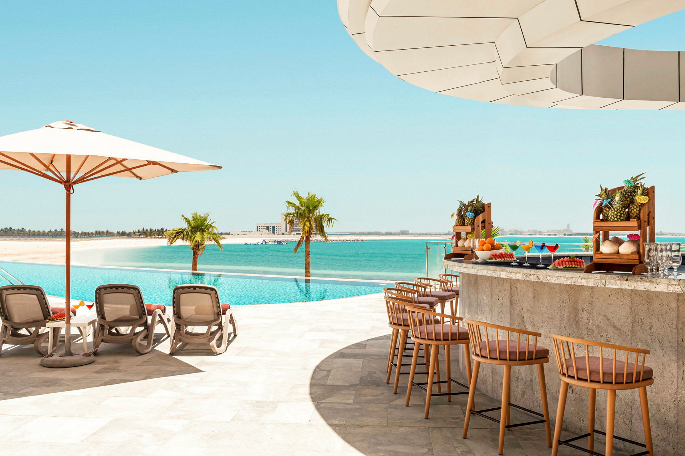 En pool ved stranden med liggestole, parasol og en poolbar med eksotiske frugter. (AI-genereret tekst)