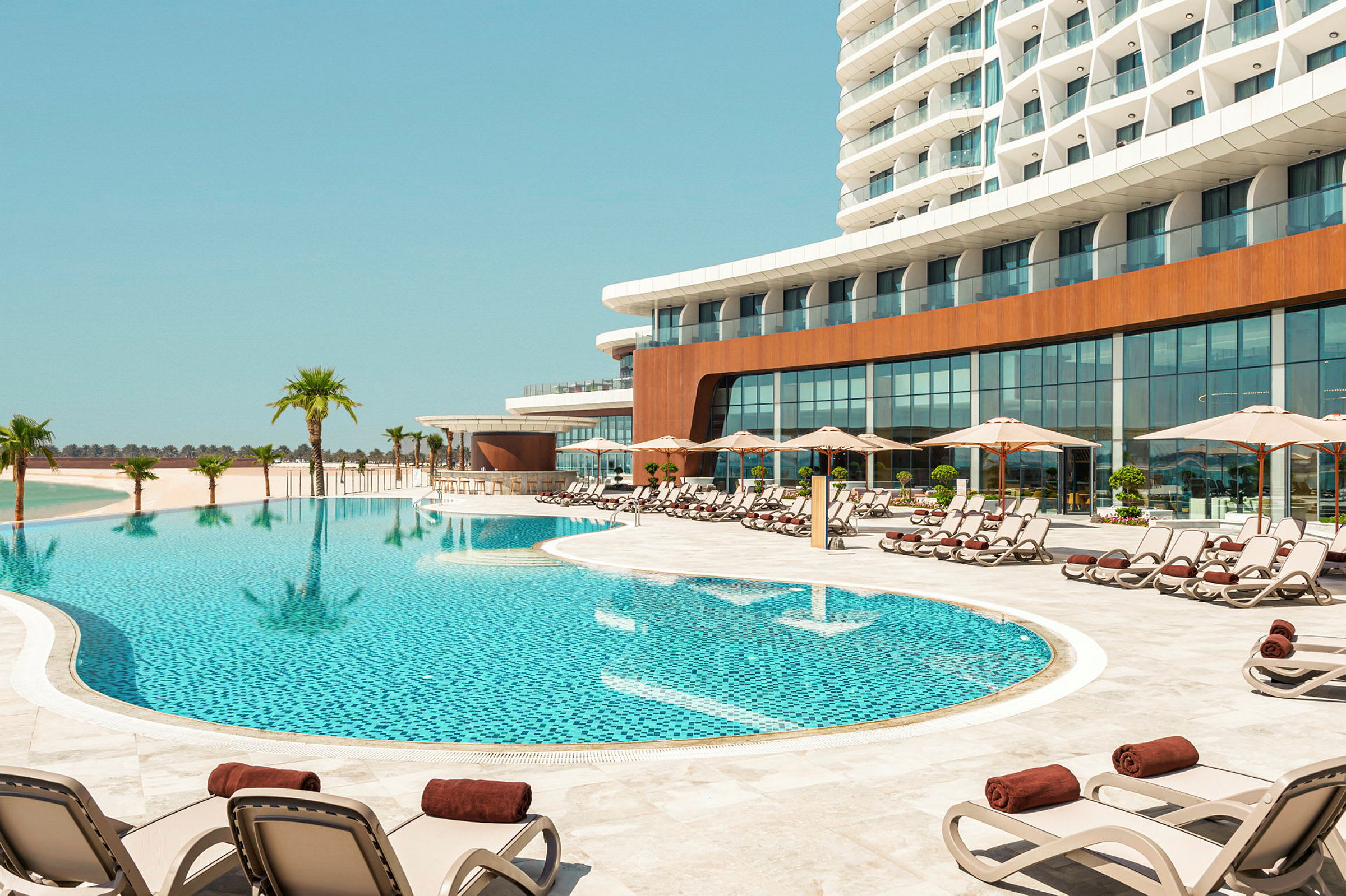 En udendørs swimmingpool omgivet af liggestole under parasoller ved en moderne hotelbygning. (AI-genereret tekst)