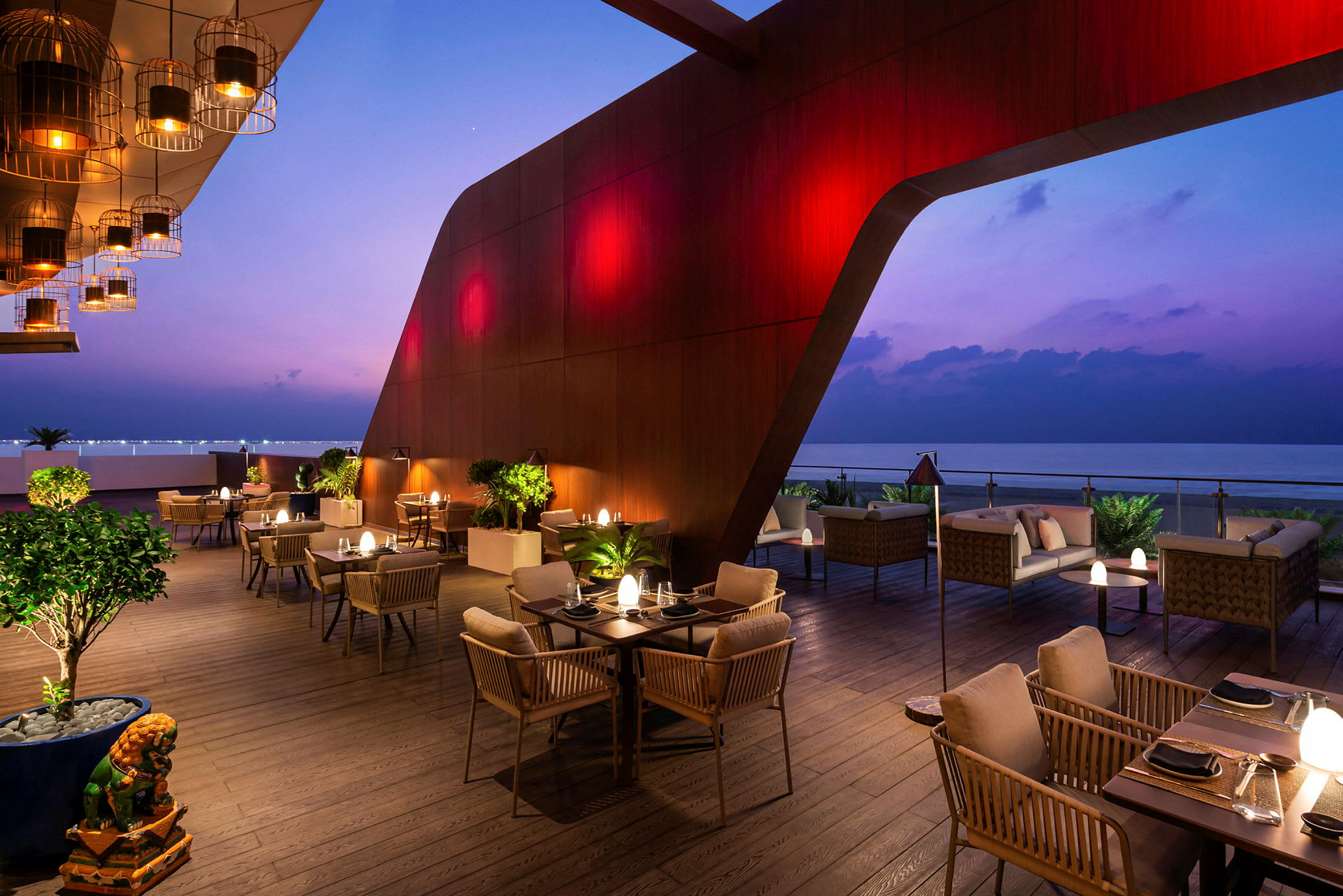 En moderne restaurantterrasse med borde og stole, beliggende ved havet om aftenen med lamper og stilfuld belysning. (AI-genereret tekst)