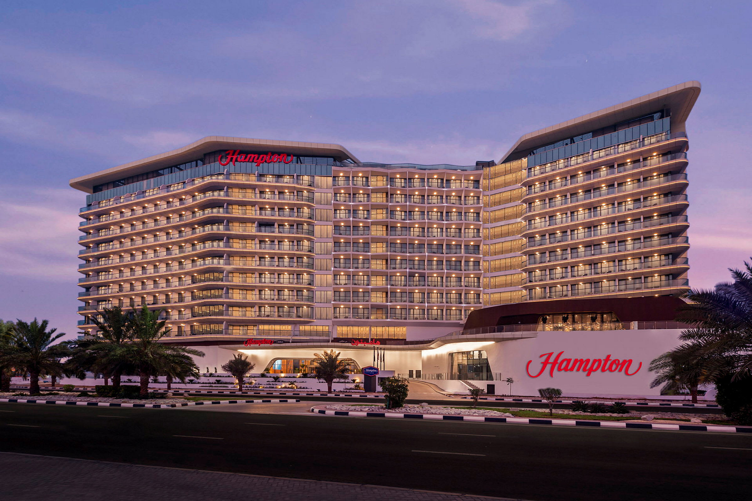 Et stort hotel med navnet "Hampton" ses i aftenbelysningen. Bygningen har flere etager med mange altaner, og der er palmer foran. (AI-genereret tekst)