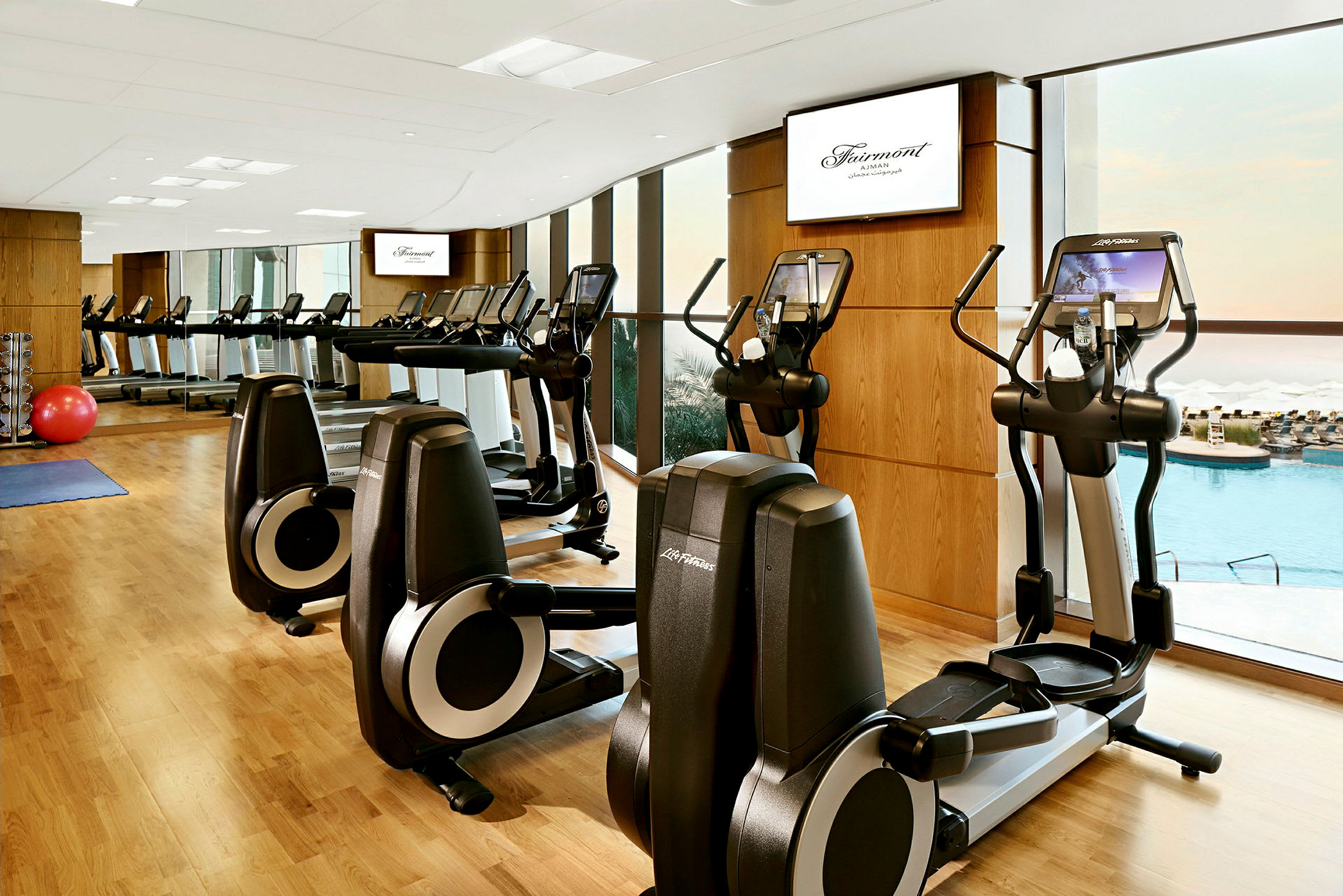 Et fitnesscenter med træningsmaskiner, trævægge, spejle og udsigt til en swimmingpool gennem store vinduer. (AI-genereret tekst)
