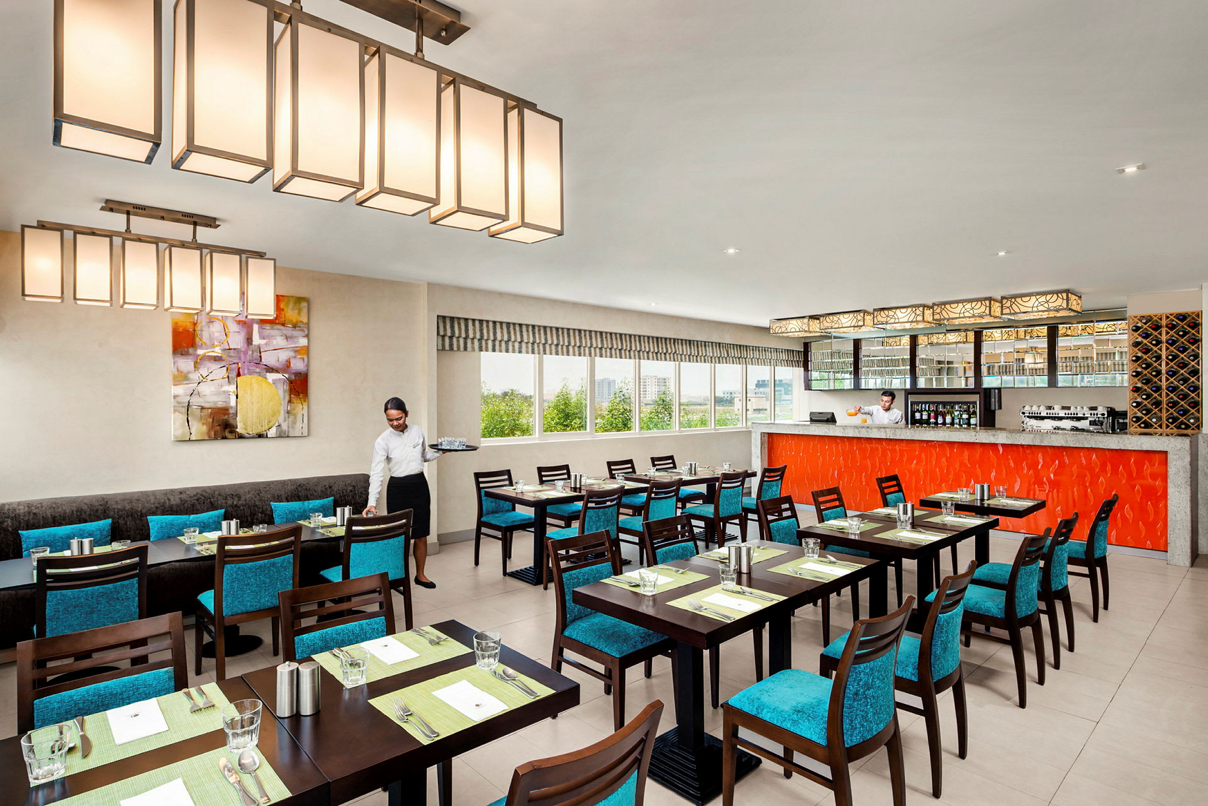 En elegant restaurant med borde og turkisblå stole. Barområdet er indrettet med en orange panel, og store vinduer giver naturligt lys. (AI-genereret tekst)