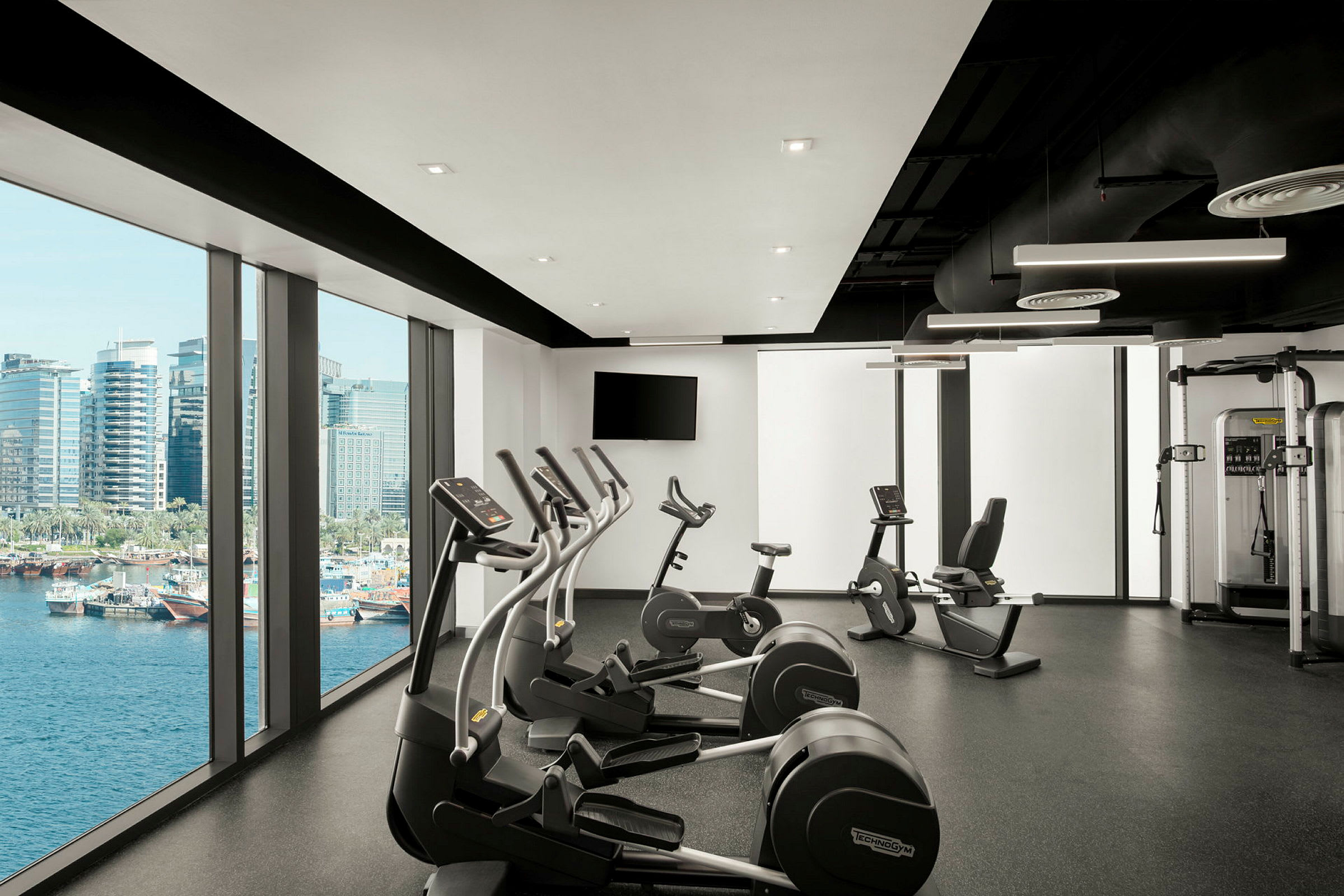 Et moderne træningscenter med fitnessmaskiner og store vinduer med udsigt over vandet og byens skyline. (AI-genereret tekst)