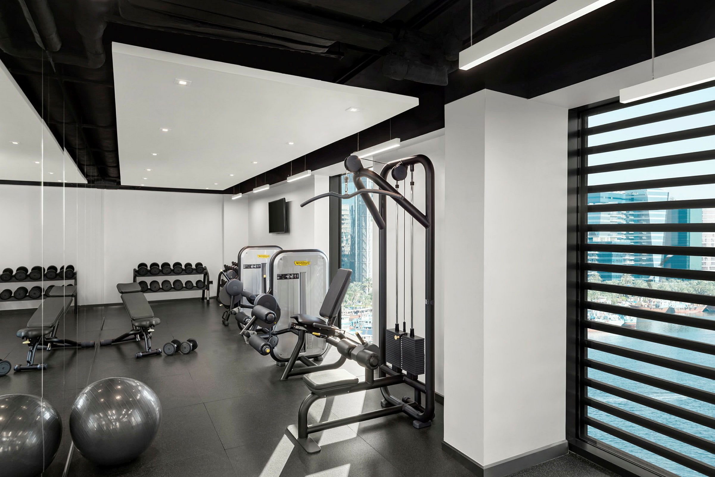 Et fitnesscenter med træningsudstyr, vægte og en bænk, med vinduer der viser en udendørs udsigt. (AI-genereret tekst)