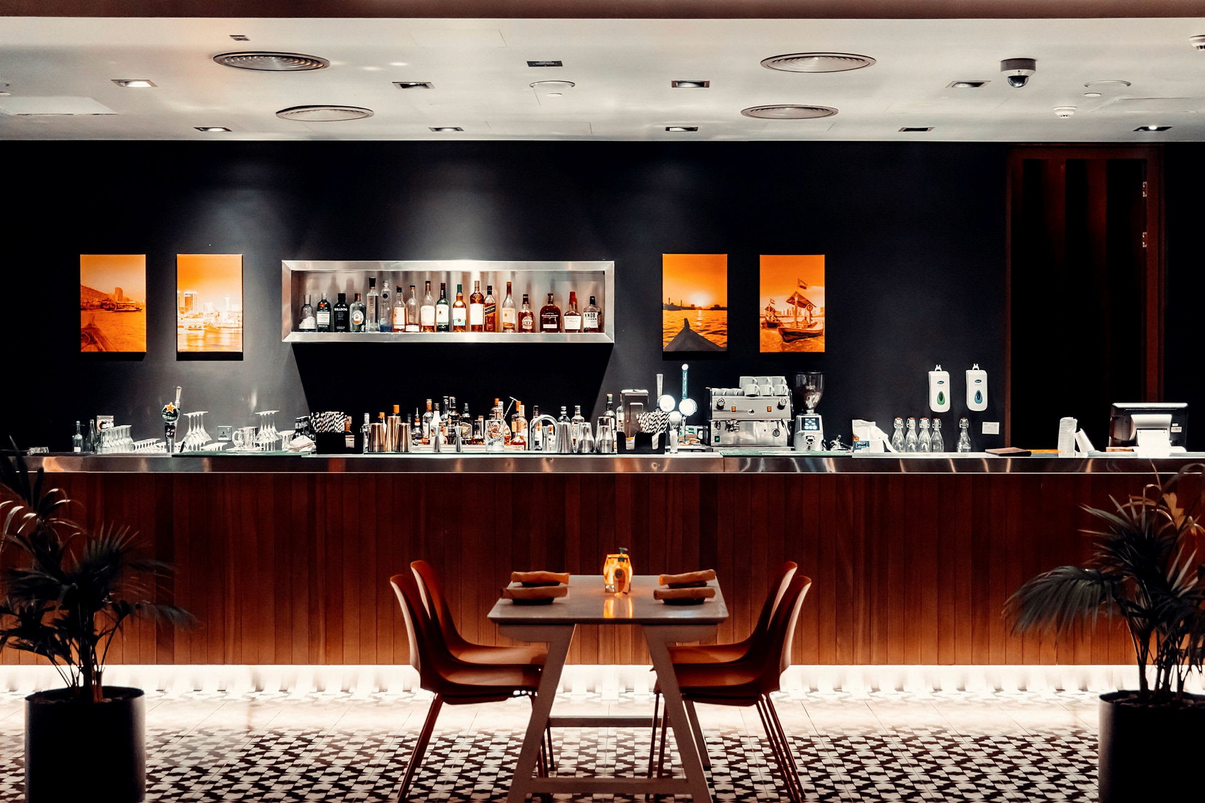 En elegant bar med hylder fyldt med flasker, en bænk med bartilbehør og et bord dækket til gæster i forgrunden. (AI-genereret tekst)
