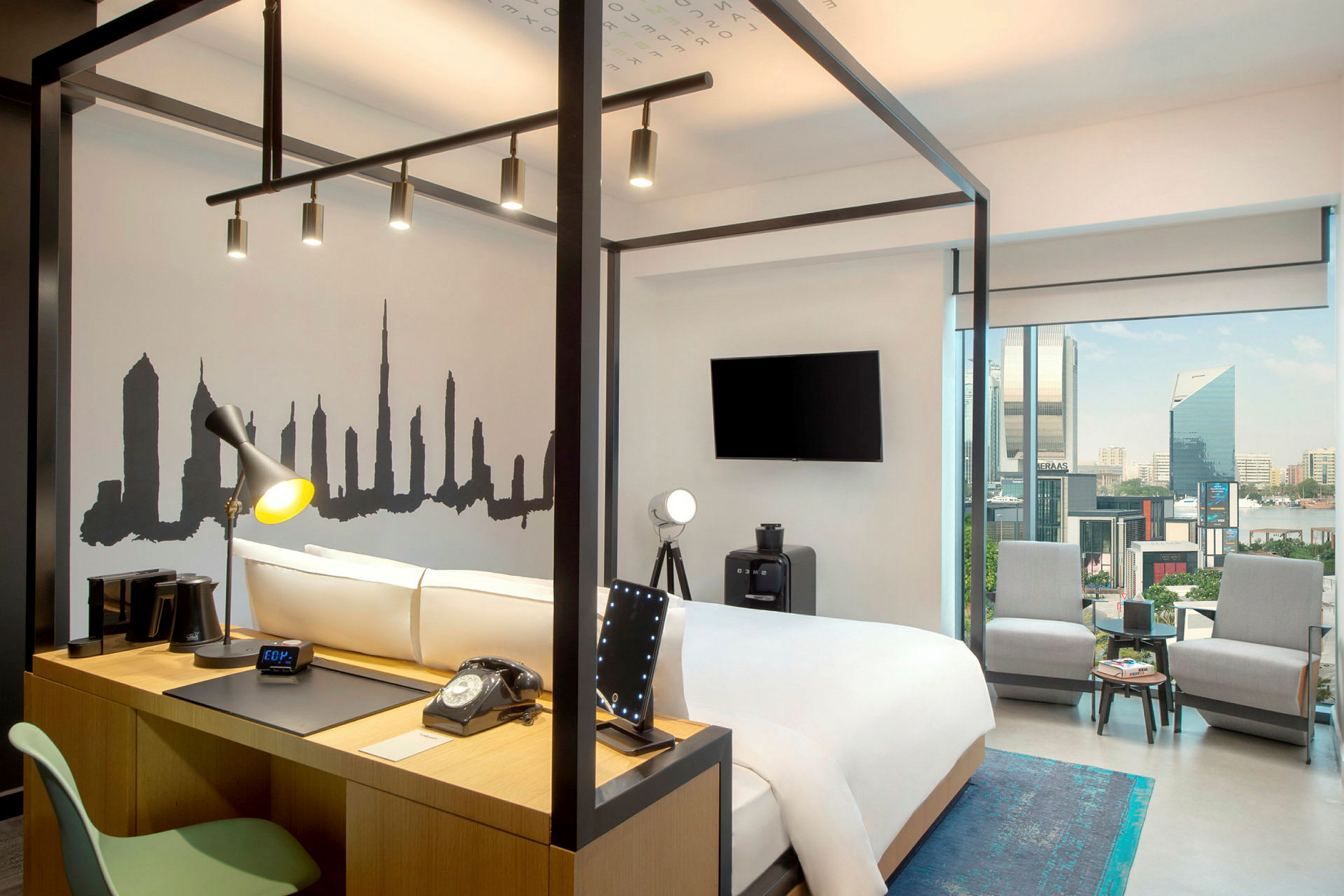 Et moderne hotelværelse med en seng omgivet af en stålkonstruktion, en vægdekoration der viser en bysilhuet, og et skrivebord med lampe og telefon. Store glasvinduer giver udsigt over byen. (AI-genereret tekst)