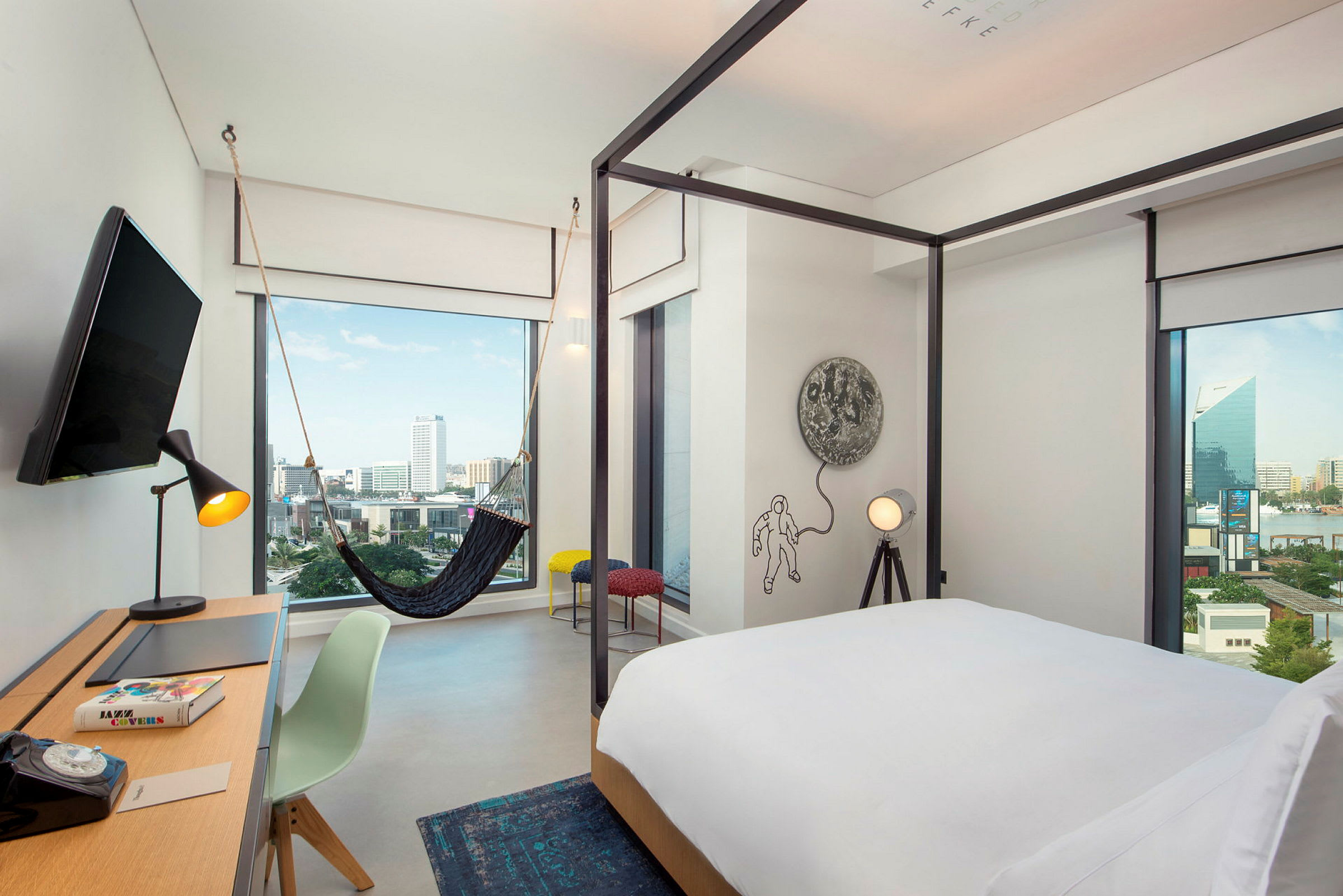 Et moderne hotelværelse med en seng med hvidt sengetæppe, et skrivebord med en stol og en lampe samt en hængekøje nær et stort vindue, der viser udsigt over byen. (AI-genereret tekst)