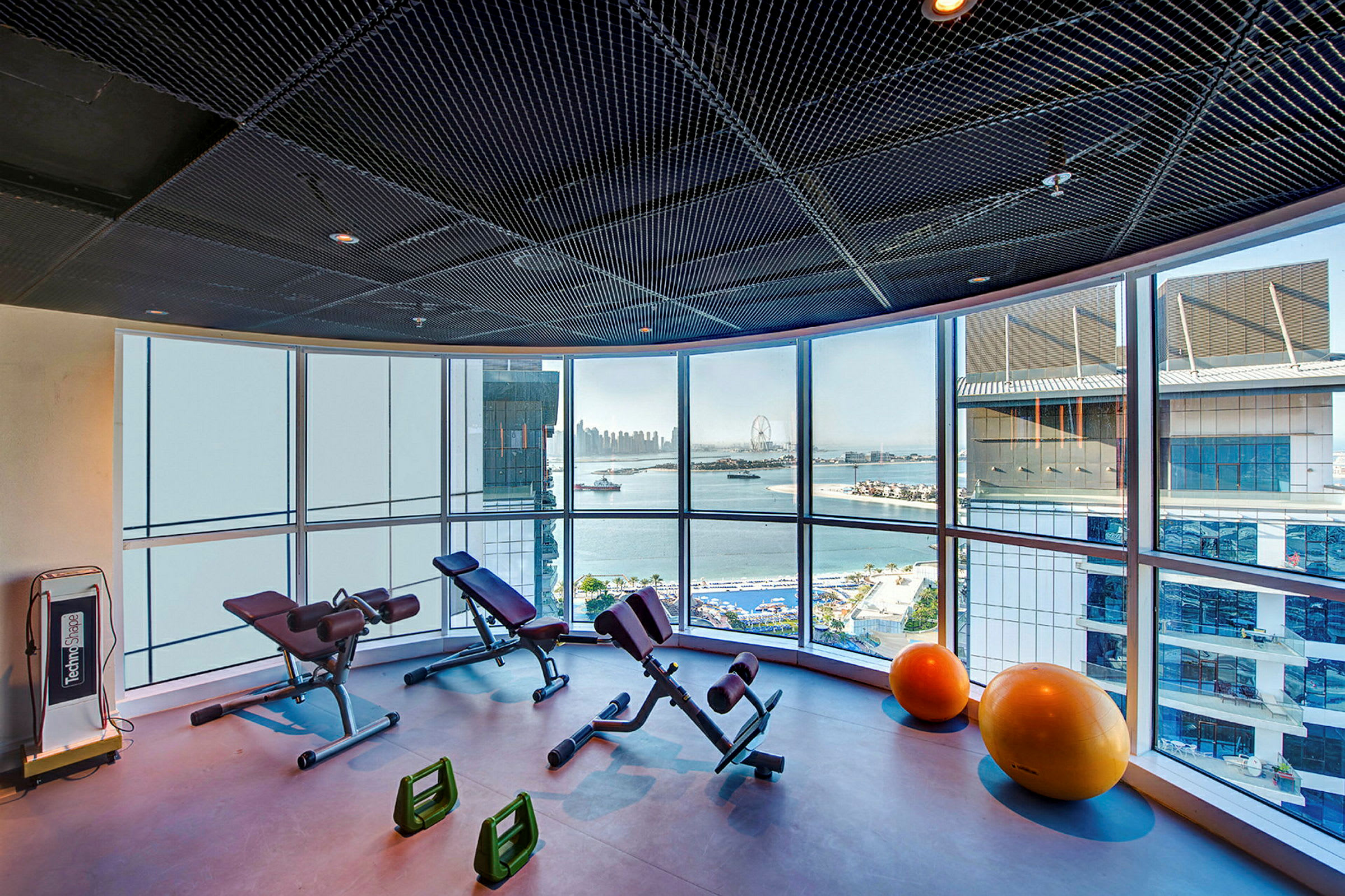 Et fitnessrum med træningsudstyr og store vinduer med udsigt over havet og bymiljø. (AI-genereret tekst)