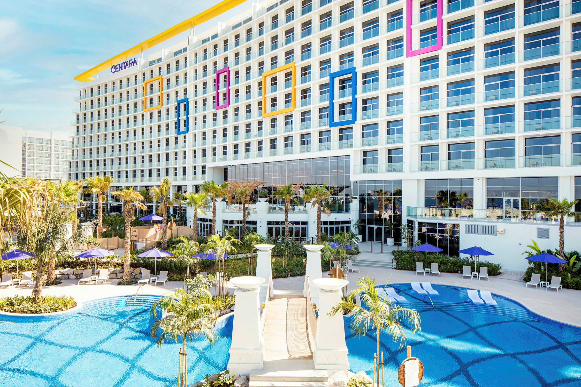 Et moderne hotel med store vinduer, omgivet af palmer og en pool med liggestole og parasoller. (AI-genereret tekst)