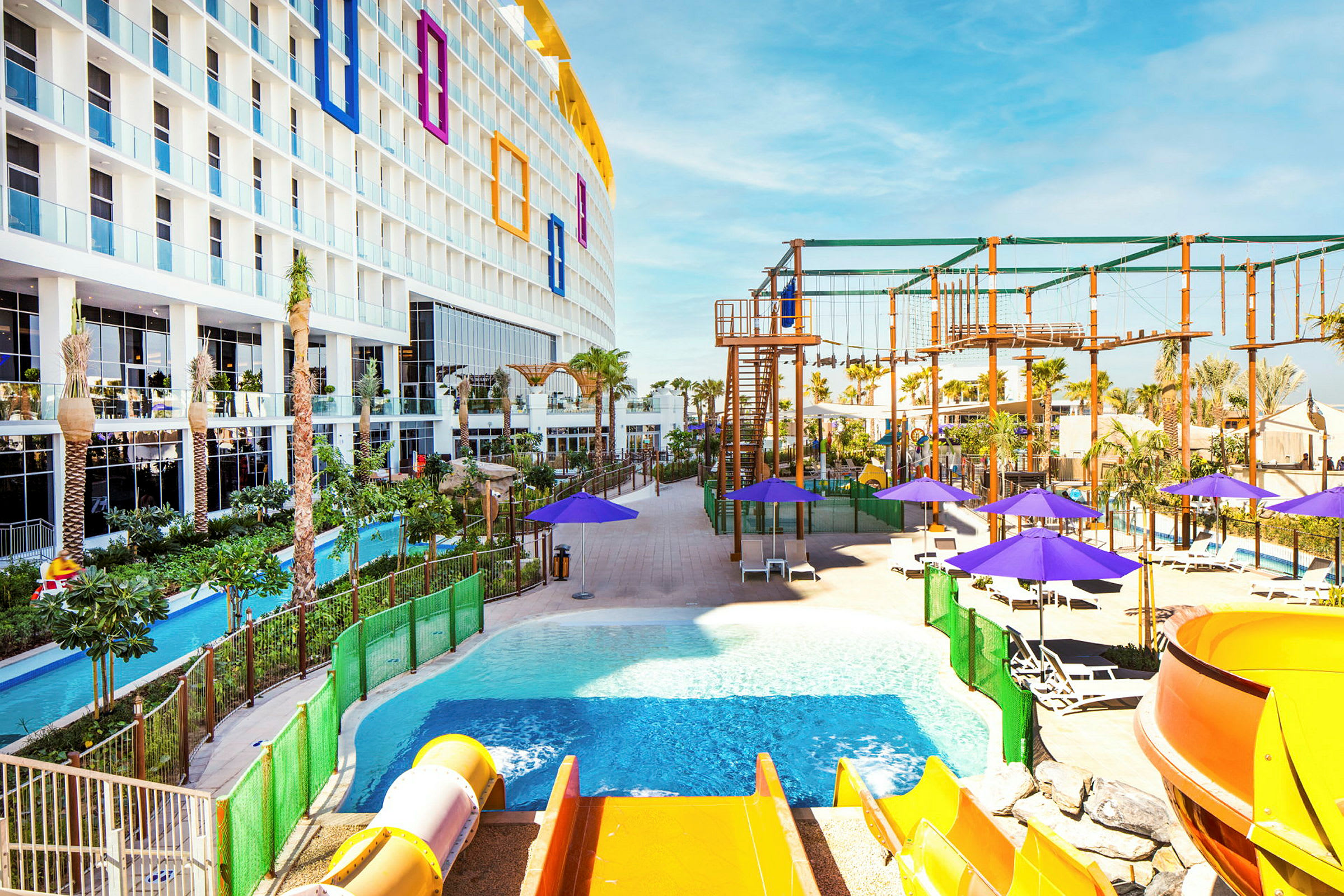 Et hotel med flere etager og altaner med udsigt over et poolområde med en vandrutsjebane, liggestole og parasoller. I baggrunden er der et klatreområde og beplantning. (AI-genereret tekst)