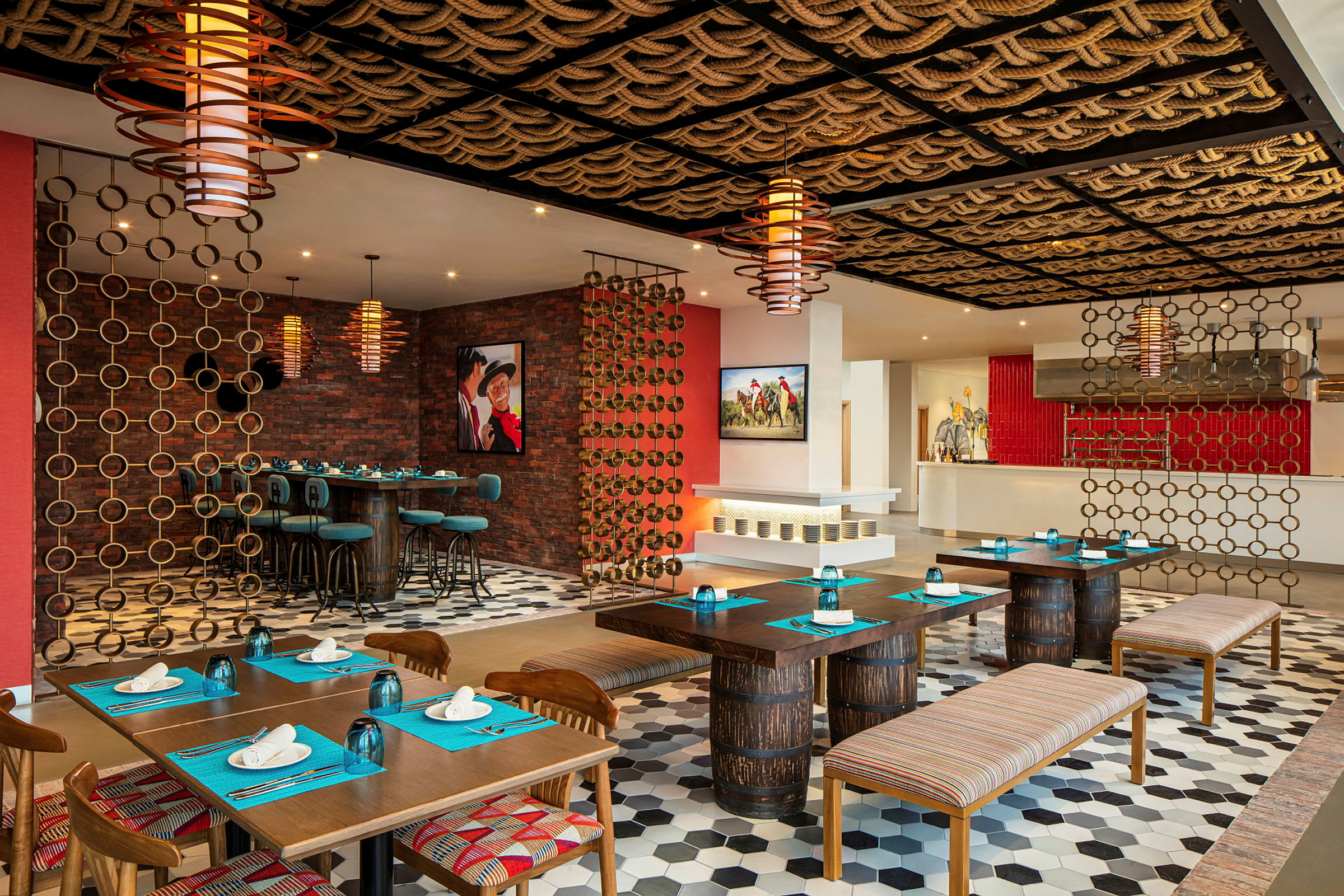 Et indrettet restaurantområde med borde og stole i træ. Dekorationer inkluderer reb i loftet og metalringe. Farverne går i jordtoner med indslag af rødt og blåt. (AI-genereret tekst)