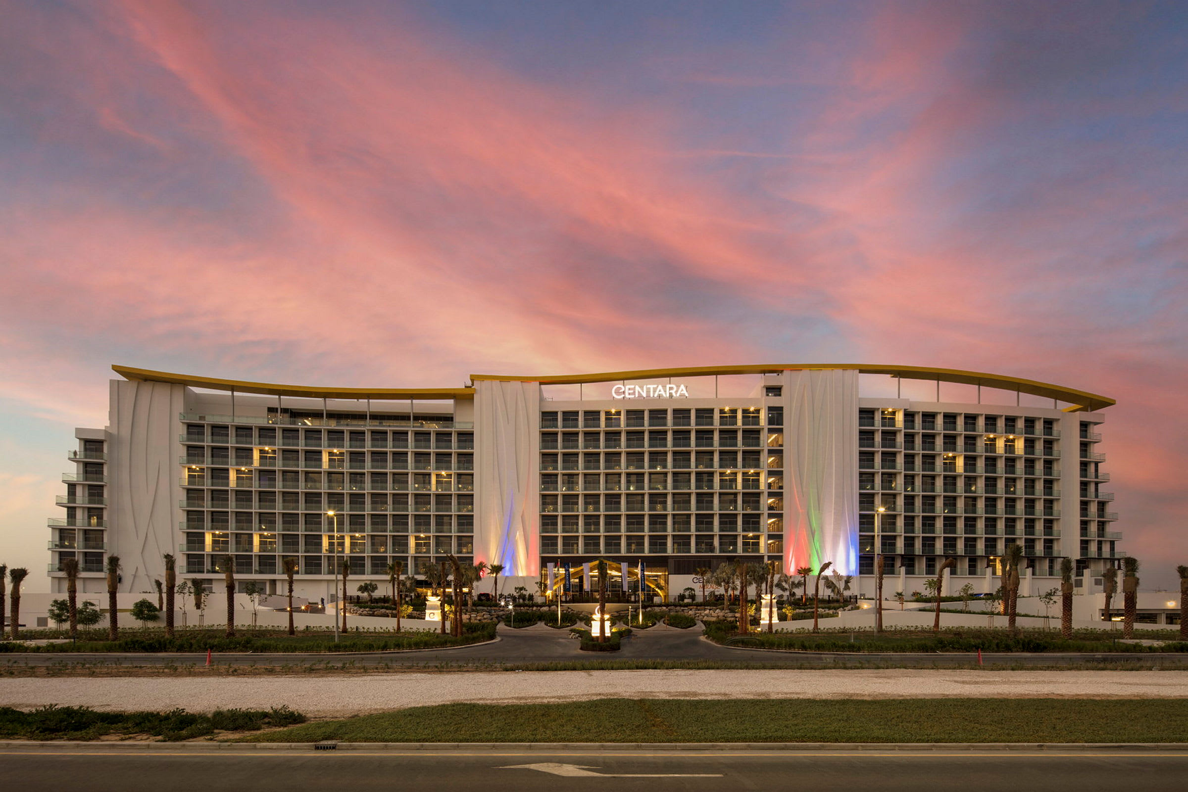 Et stort hotel med flere etager og en moderne facade, oplyst af farvede lys, med en pink og blå himmel i baggrunden. (AI-genereret tekst)