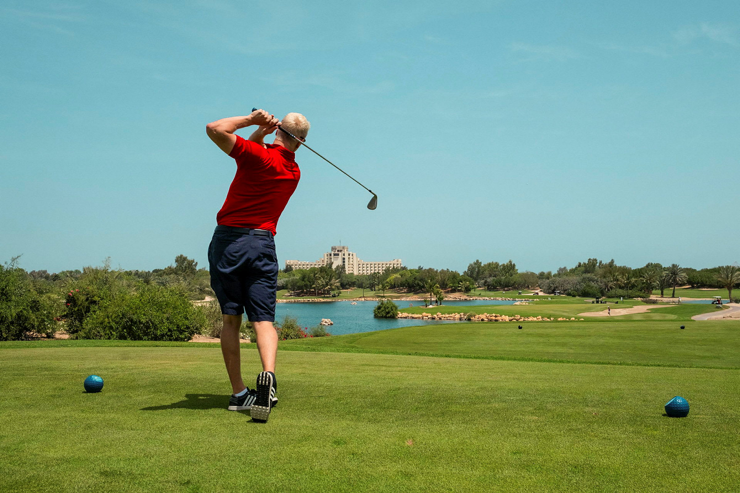 En person klædt i rødt spiller golf på en bane med grøn græsplæne, omgivet af træer og vand. Et hotel ses i baggrunden. (AI-genereret tekst)