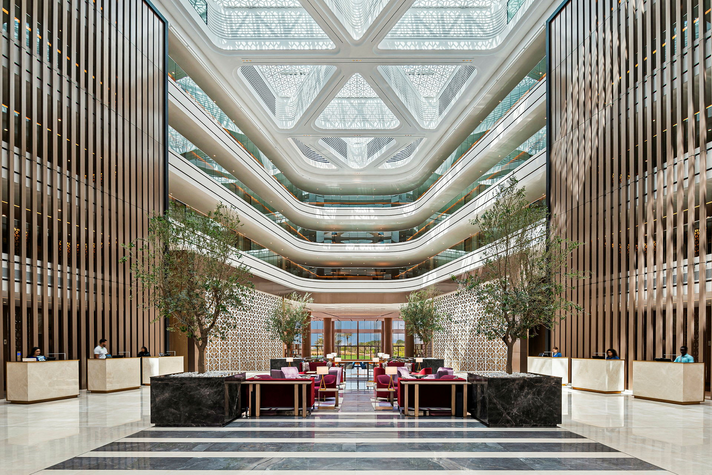 Billede af en moderne hotellobby med elegante siddeområder, frodige træer, receptionsskranker og arkitektoniske detaljer. (AI-genereret tekst)