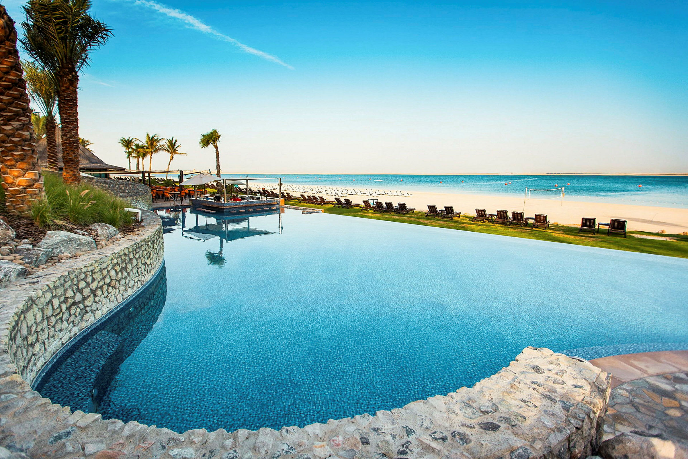 En infinitypool nær en strand med liggestole og parasoller ved det klare blå hav. (AI-genereret tekst)