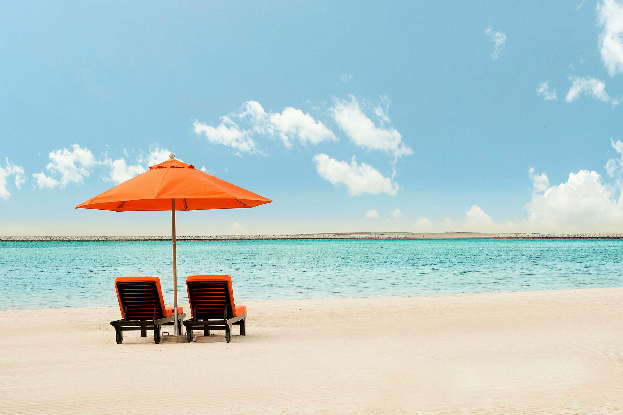 To liggestole med orange hynder står under en stor orange parasol på en sandstrand med hav i baggrunden. (AI-genereret tekst)