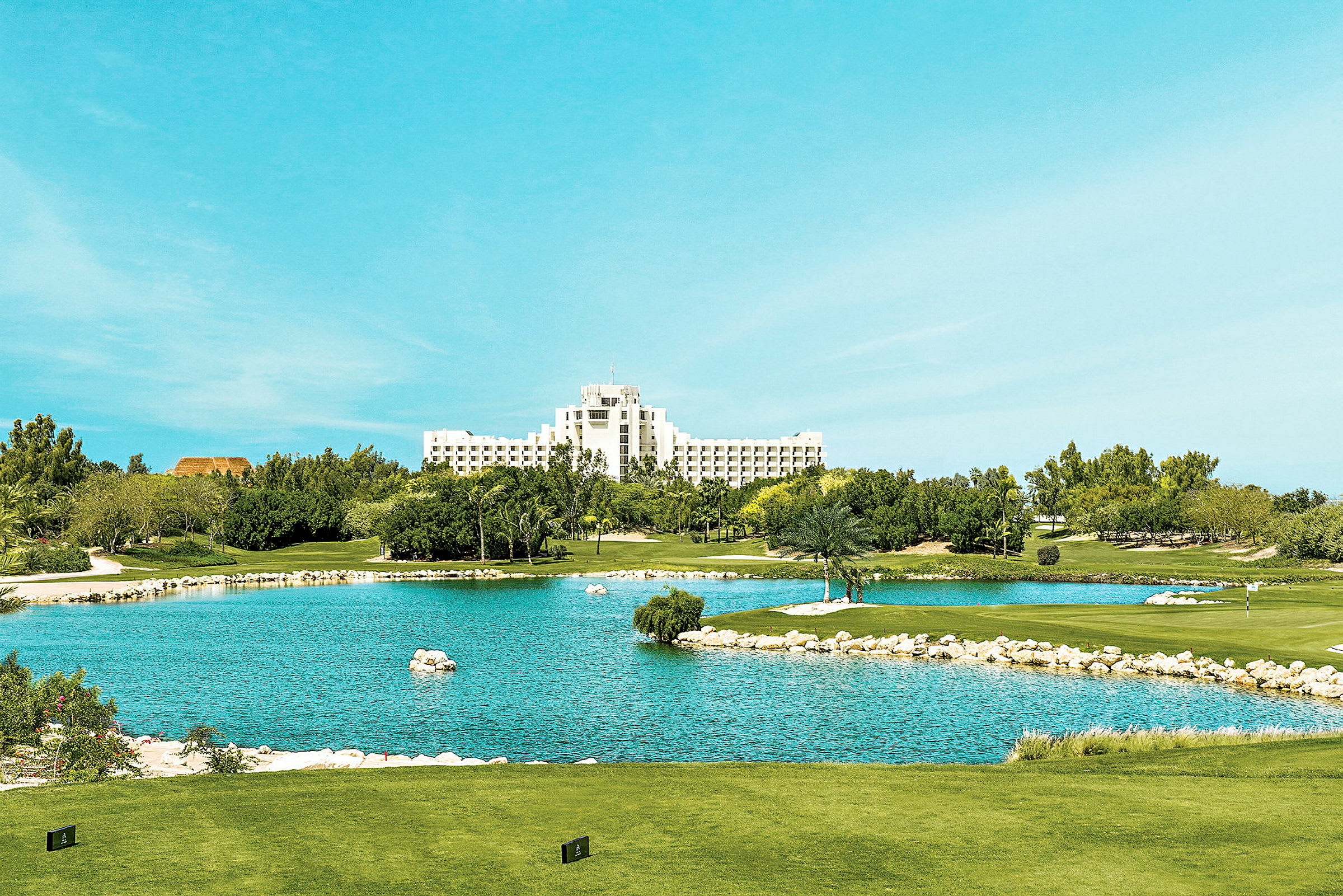 Udsigt over en golfbane med en dam i forgrunden og et stort hvidt hotel i baggrunden. (AI-genereret tekst)