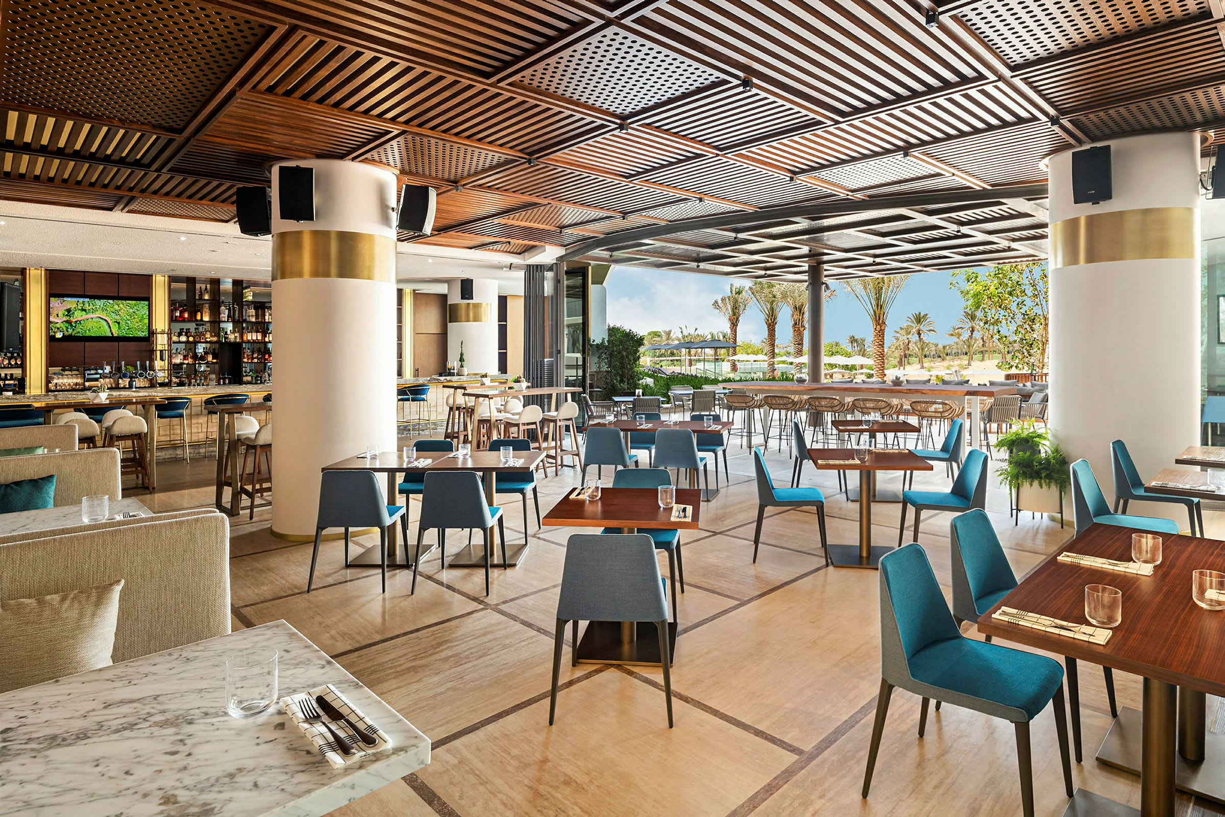 Restaurant med moderne borde og stole samt åbning mod terrasse med udsigt over palmer og grønne områder. (AI-genereret tekst)