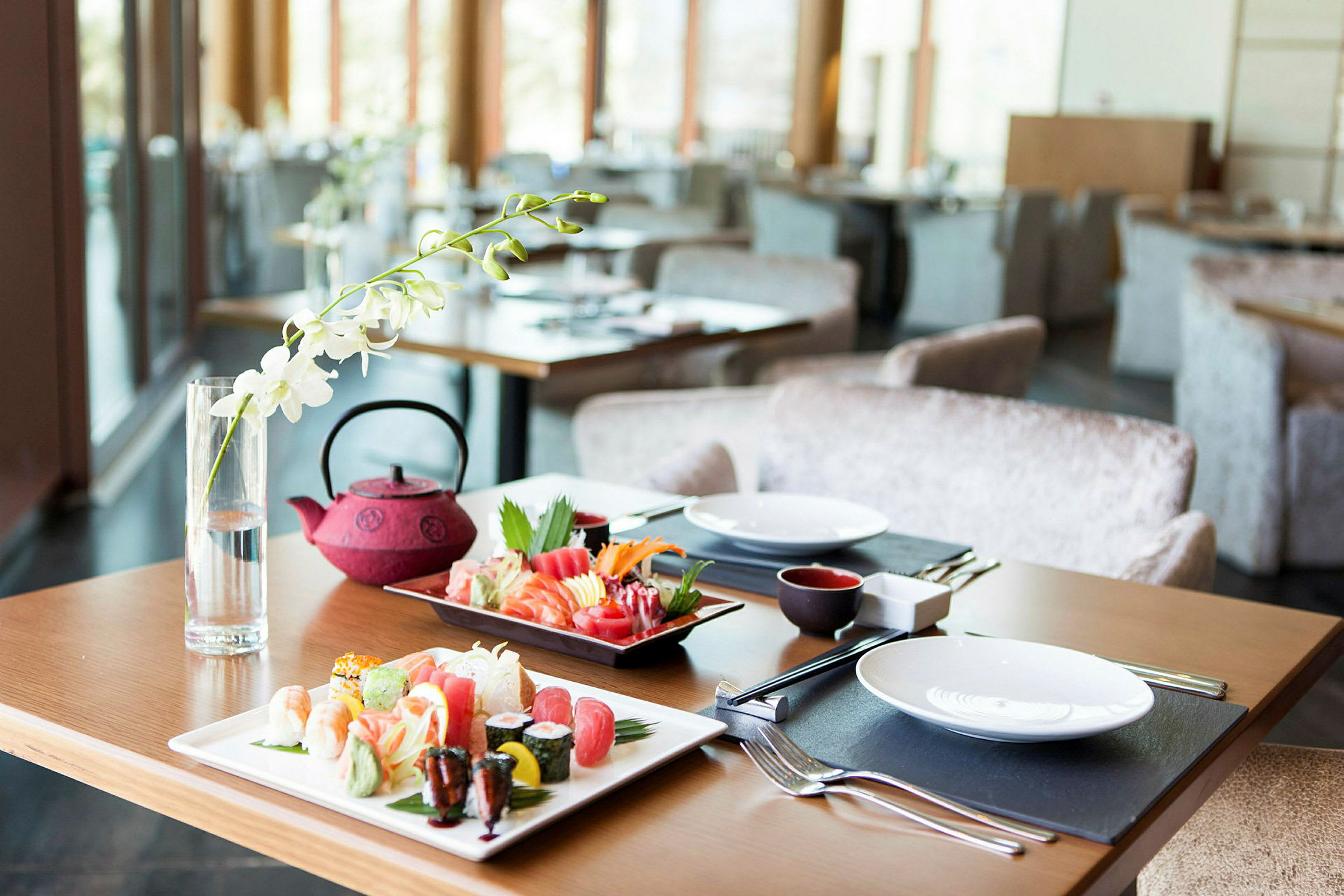 Et bord på en restaurant er dækket med sushi på fade, en rød tekande og et glas med en hvid blomst som dekoration. (AI-genereret tekst)