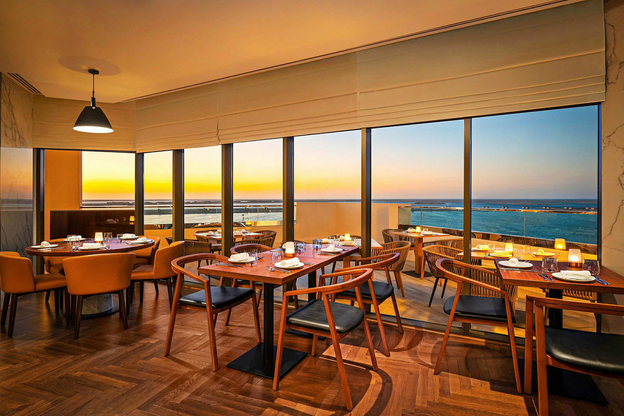 Interiør af en restaurant med borde og stole samt udsigt over havet i solnedgang. (AI-genereret tekst)