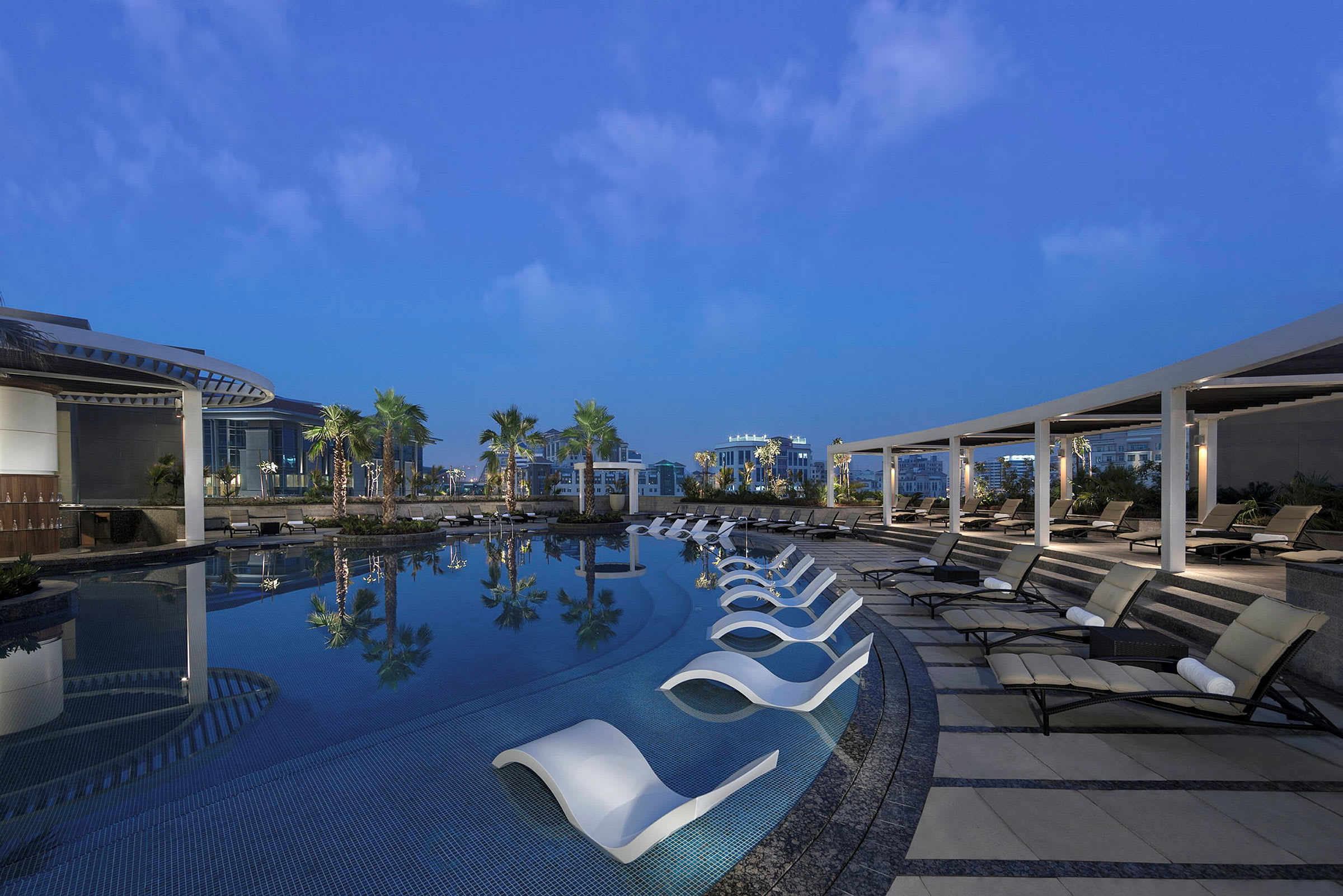 Udendørs pool med liggestole ved siden af, omgivet af palmer og bygninger om aftenen. (AI-genereret tekst)