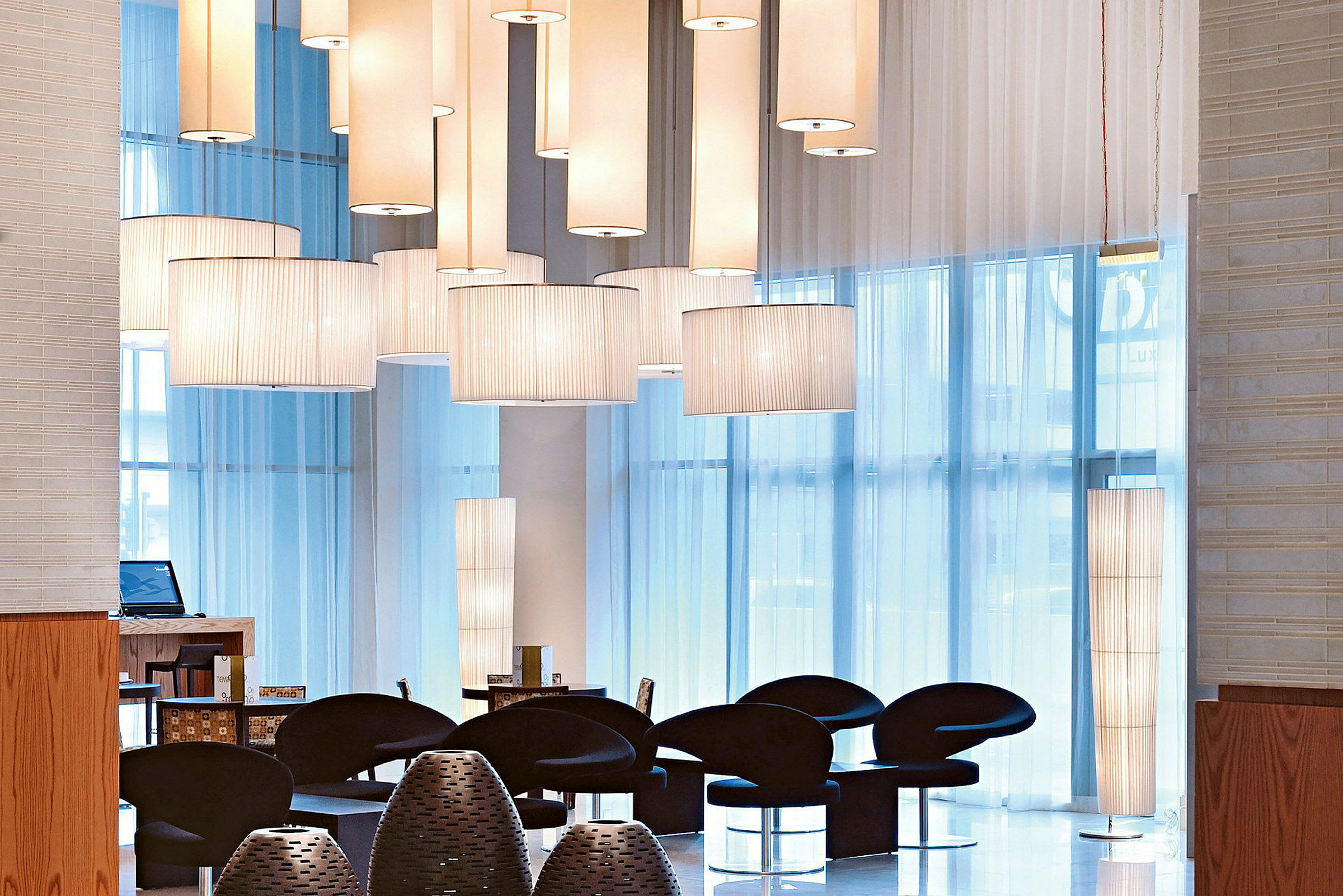 Interiørudsigt med en moderne lounge med sorte stole, hængende lamper og store vinduer, der lukker dagslys ind. (AI-genereret tekst)