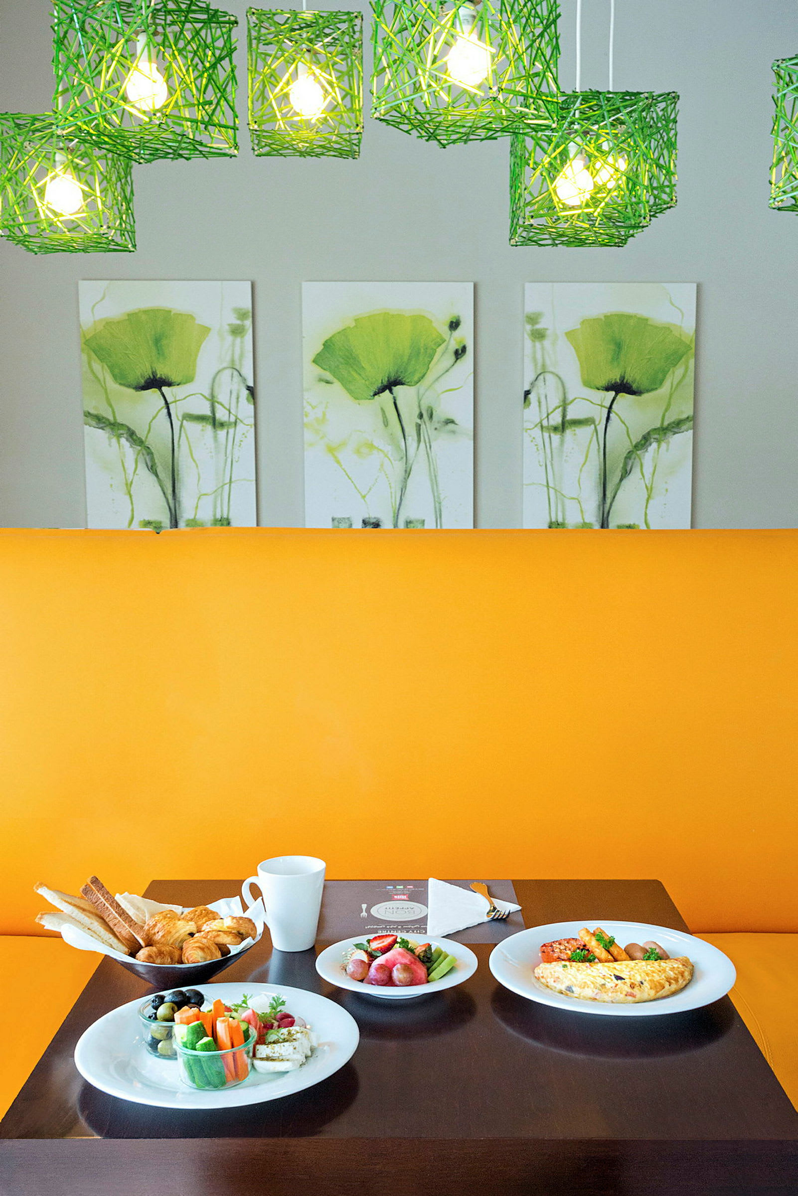 Et bord fyldt med morgenmad, herunder brød, grøntsager og omelet, foran et gult sæde med grønne lamper og blomsterkunst i baggrunden. (AI-genereret tekst)